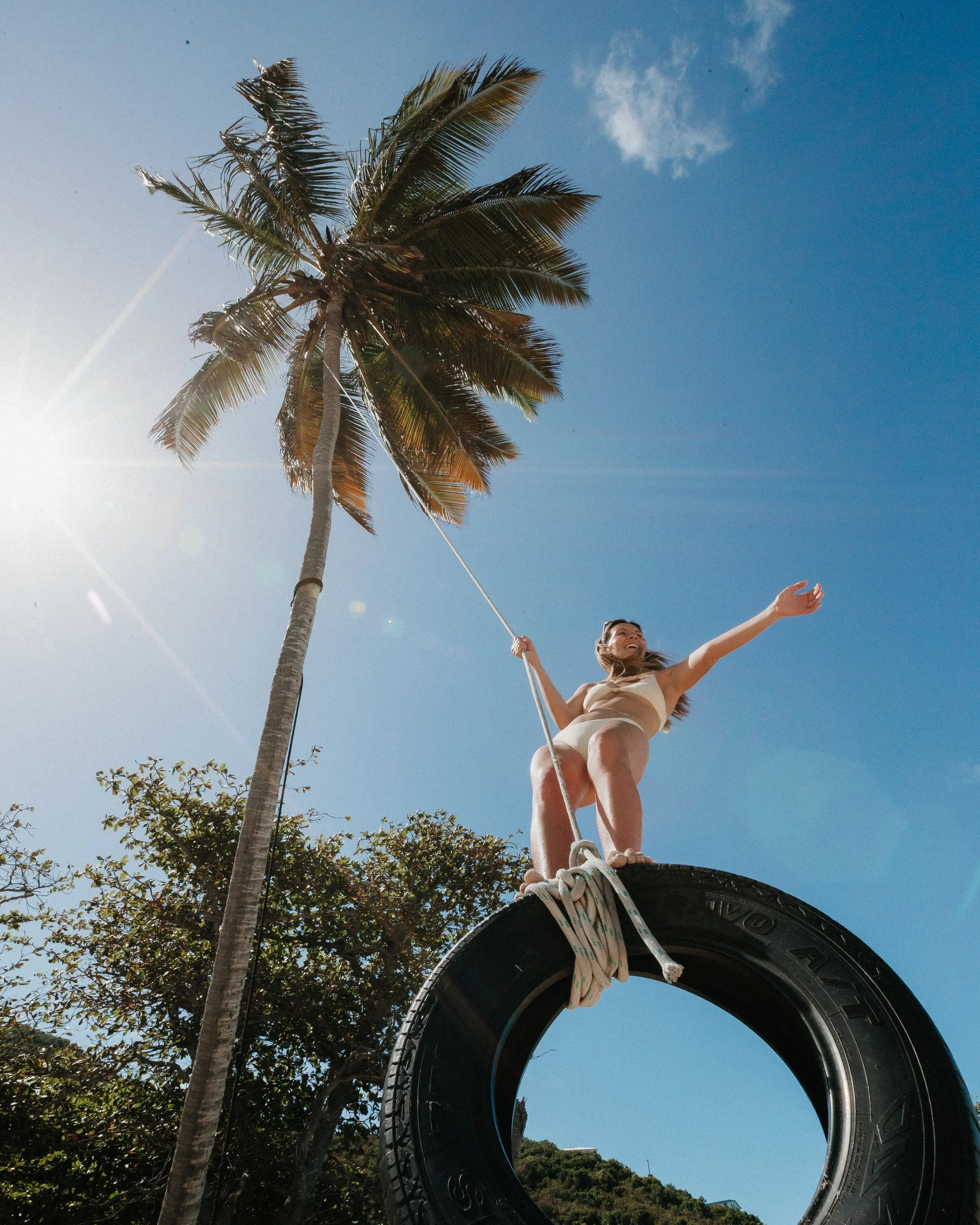A young woman riding a tire swing tied to a palm tree against a blue sky with some clouds, with a hillside and house in the distance.