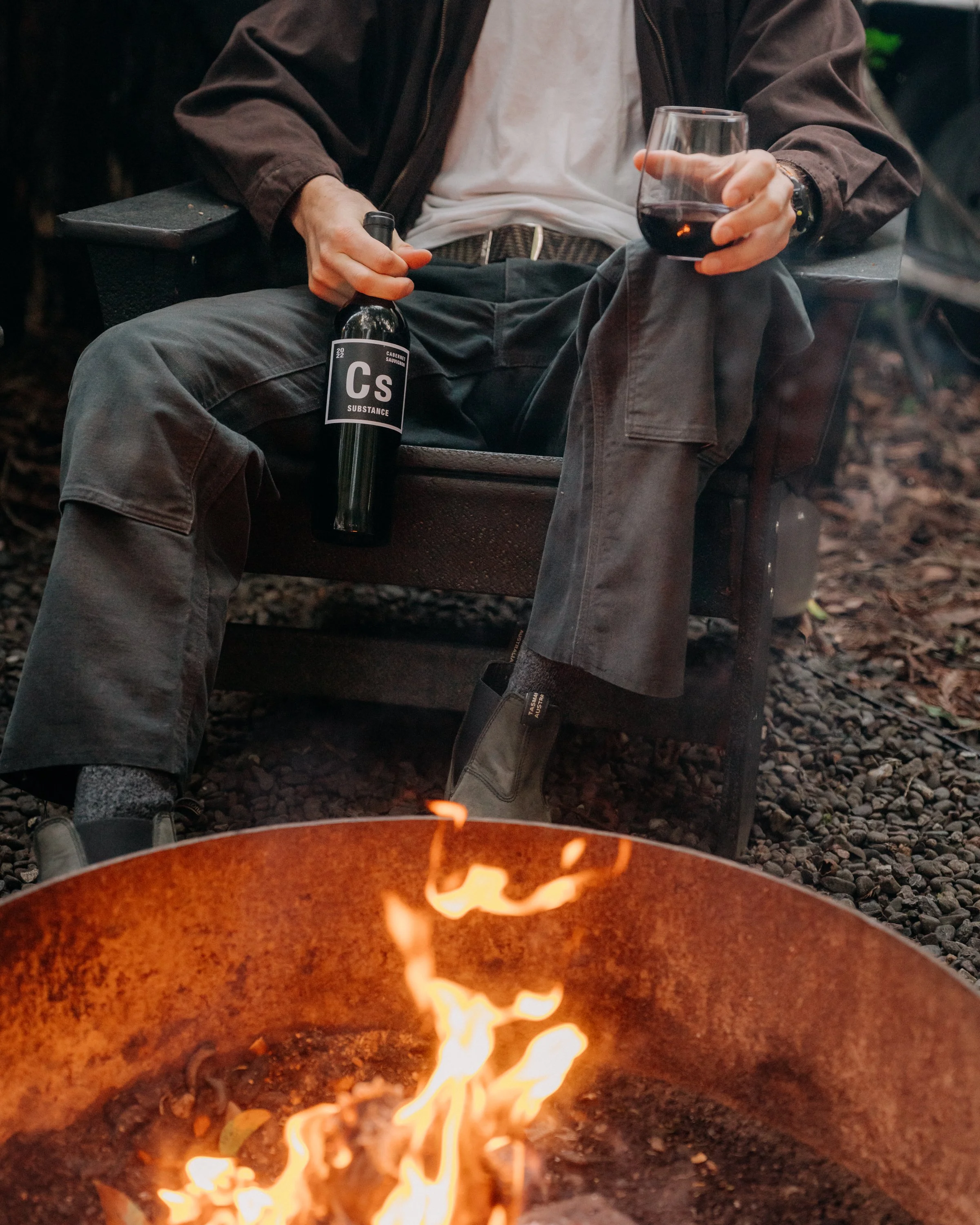 A person sitting on a bench outdoors holding a glass of red wine in one hand and a wine bottle in the other, with a fire burning nearby.