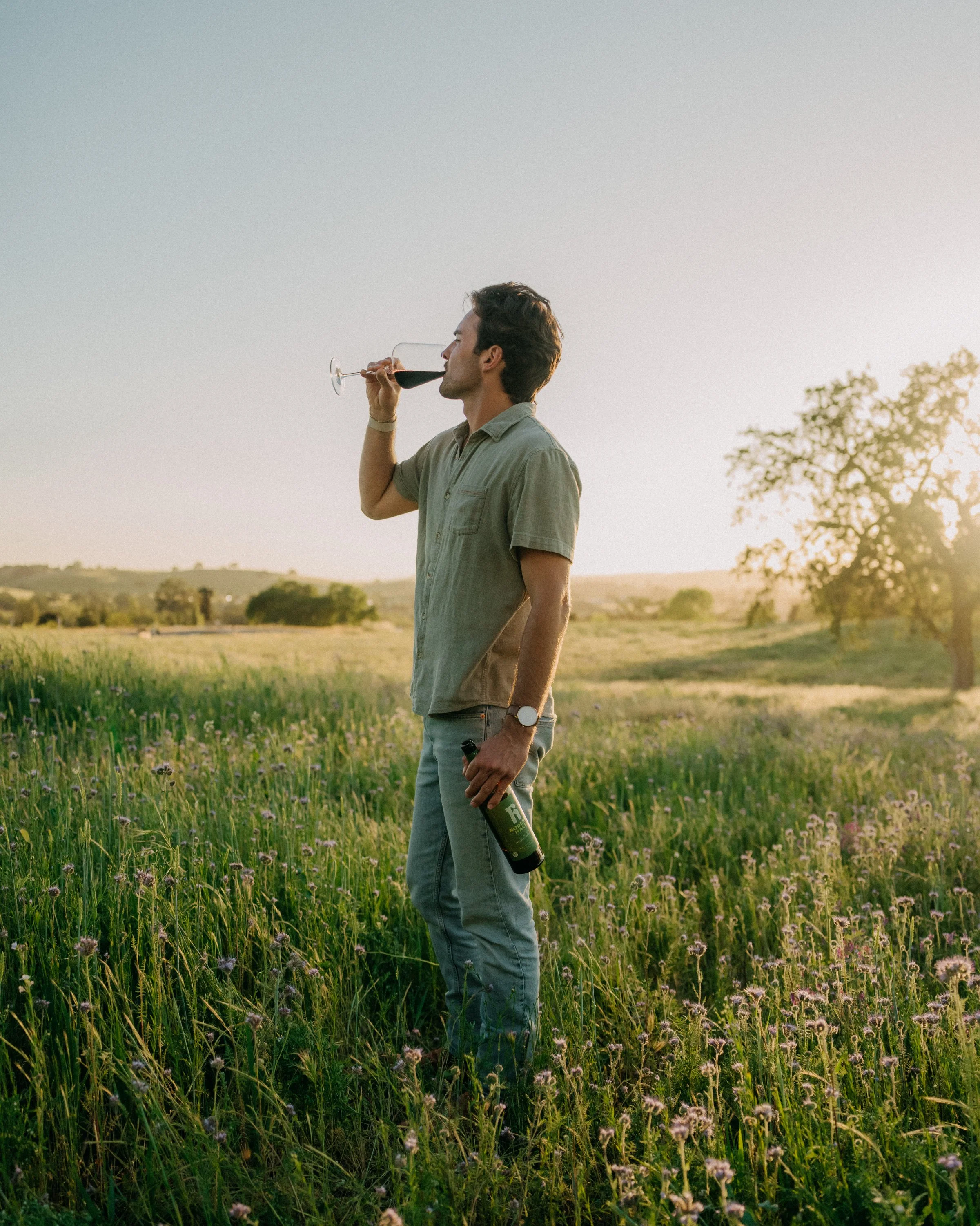A man standing in a field of tall grass and wildflowers, holding a glass of red wine and a wine bottle, with trees and a clear sky in the background during sunset.