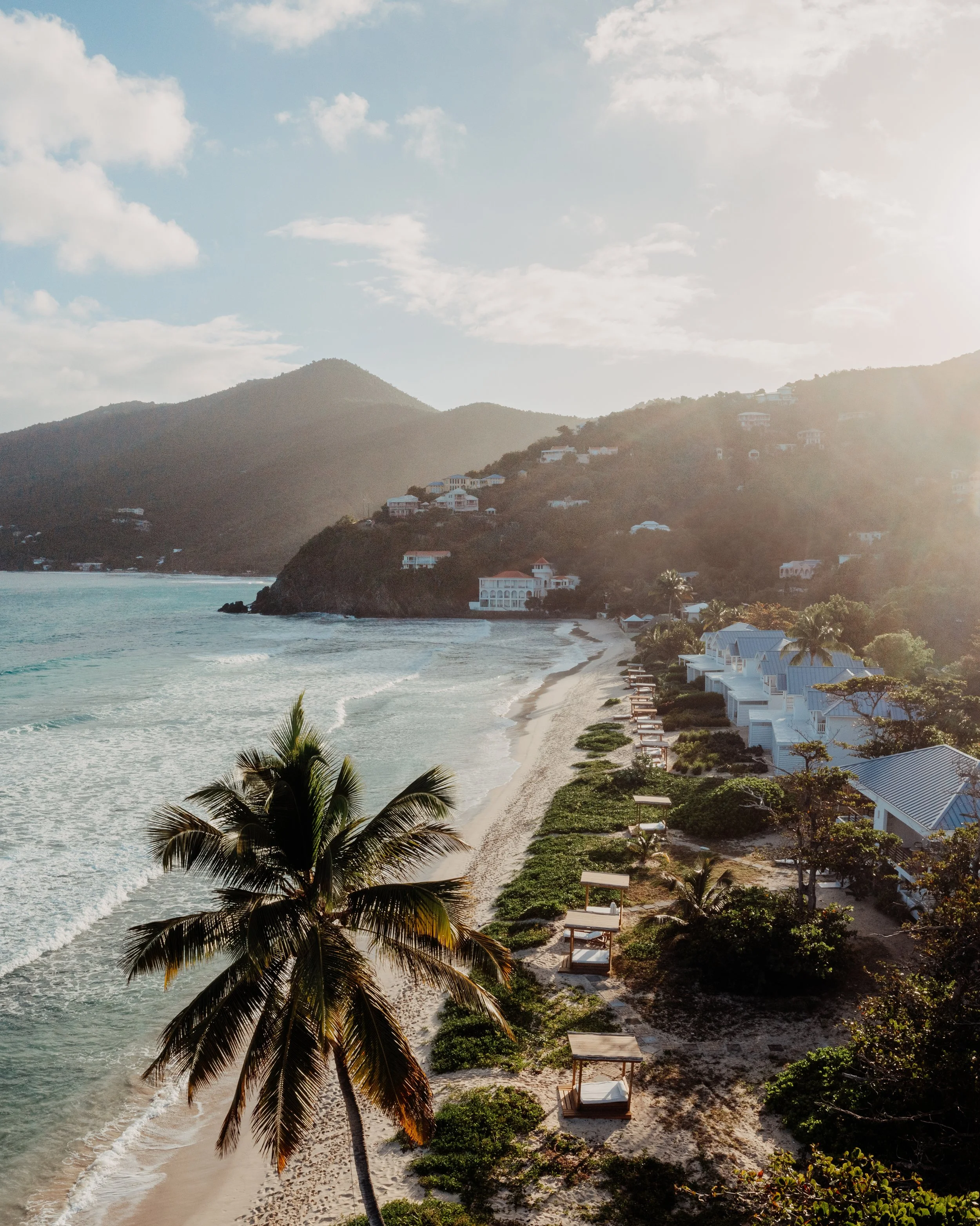 A tropical beach scene with a sandy shoreline, palm trees, beachfront cottages, and hills in the background during sunlight.