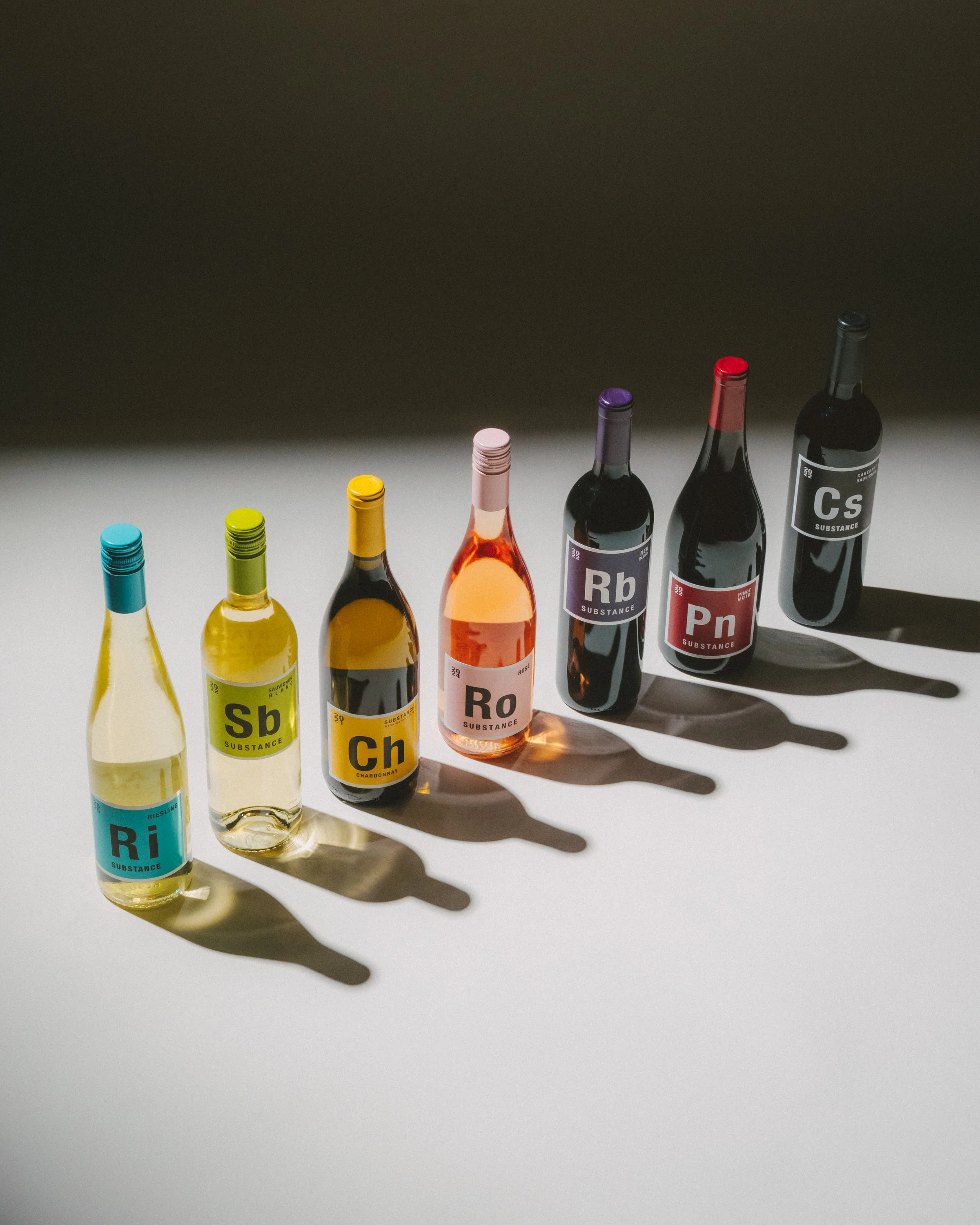Row of seven wine bottles on a white surface, casting shadows, with some bottles labeled with chemical element symbols and names.