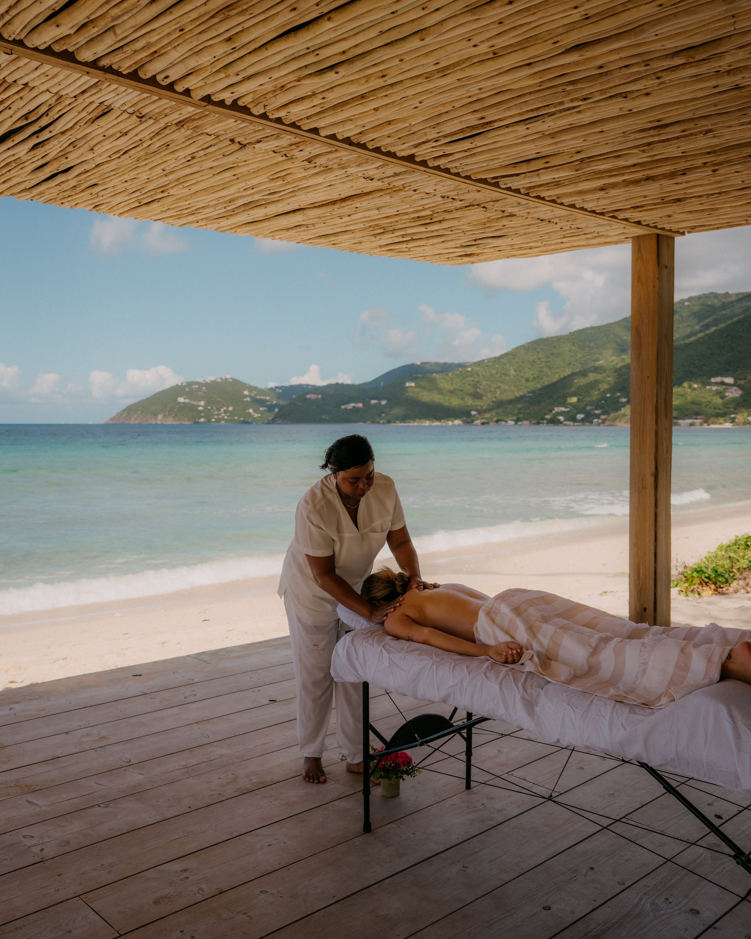 A woman receives a massage on a beachside table under a bamboo shade with the ocean, hills, and sky in the background.