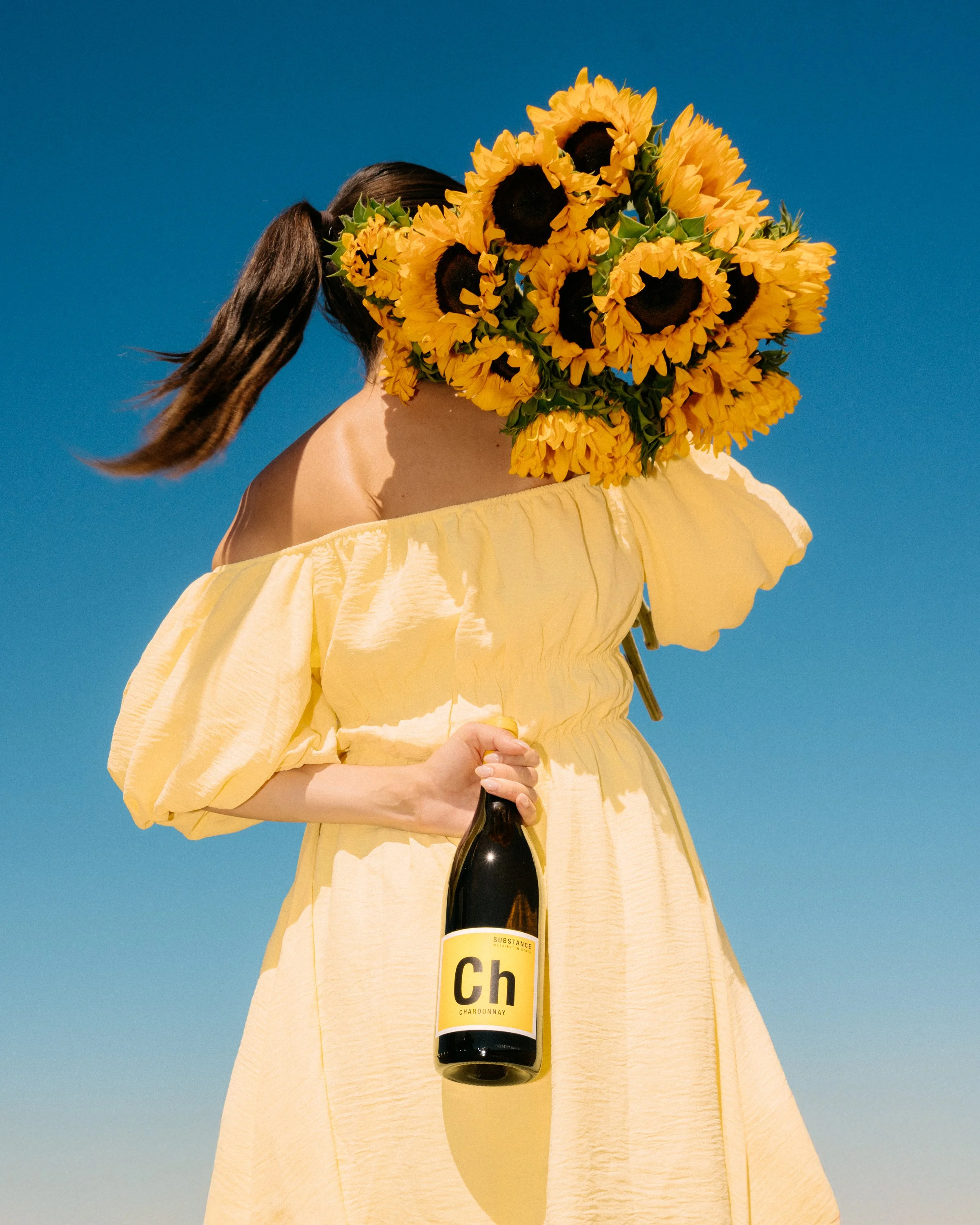A person holding a bouquet of sunflowers in front of their face, wearing a yellow off-shoulder dress, against a bright blue sky, holding a bottle of wine with a yellow label.