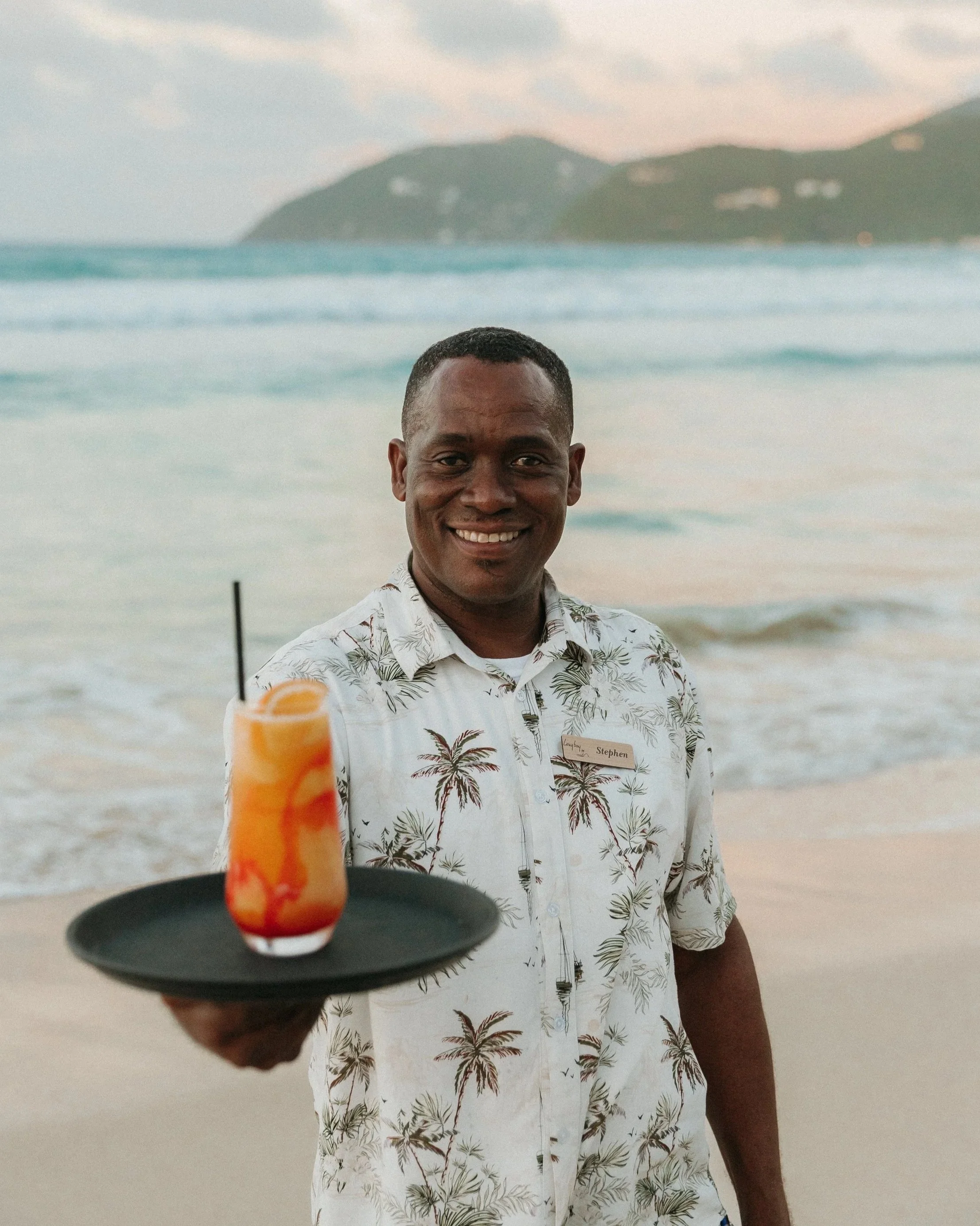 A smiling man wearing a white shirt with palm trees standing on a beach with waves and a distant island in the background, holding a tray with a tropical cocktail garnished with an orange slice.