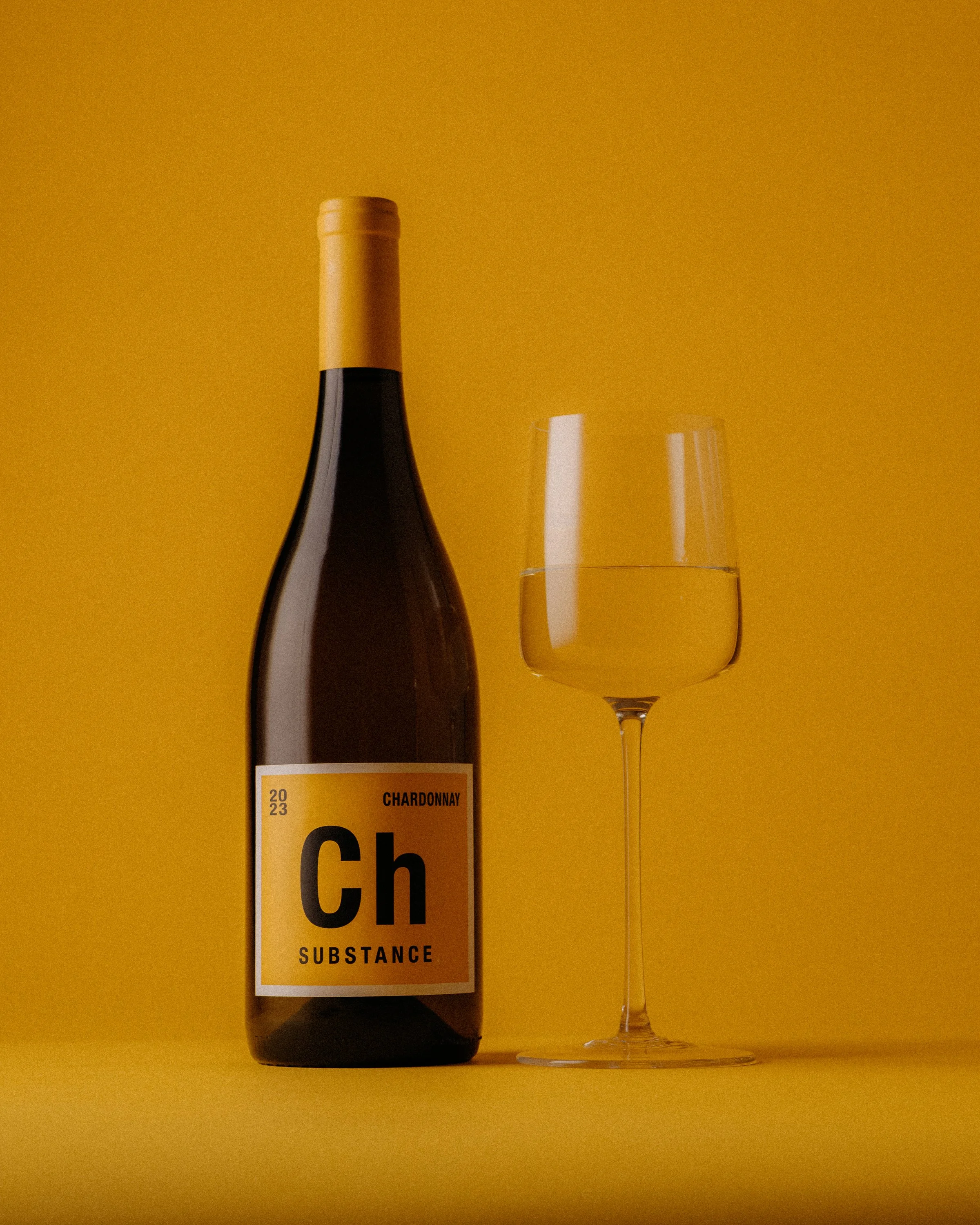 A bottle of Chardonnay wine and a wine glass filled with white wine against a yellow background.
