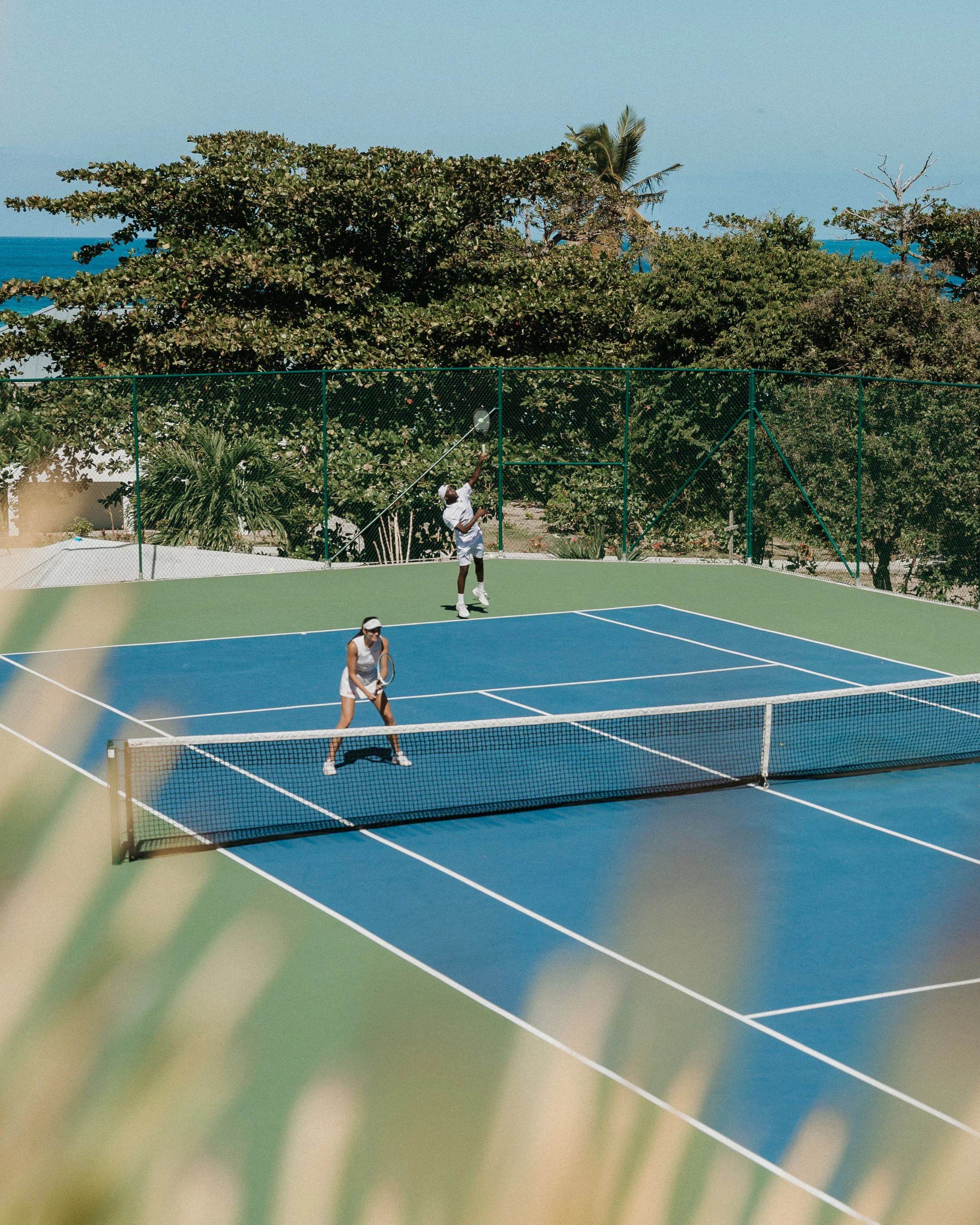 People playing tennis on an outdoor court with a view of trees and ocean in the background.