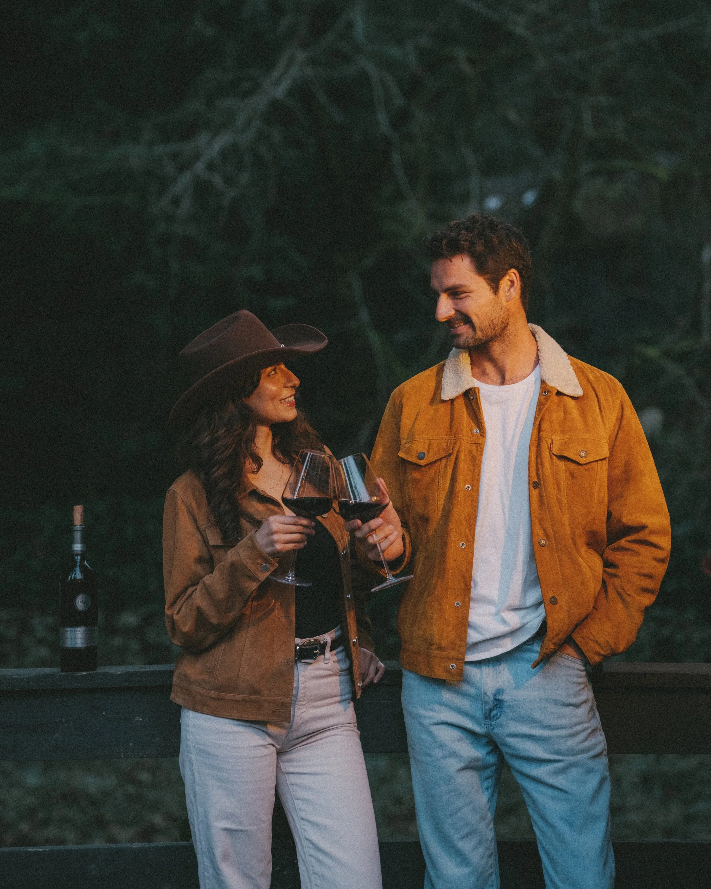 A man and a woman sharing a toast with glasses of red wine outdoors at night, smiling at each other, with trees in the background.