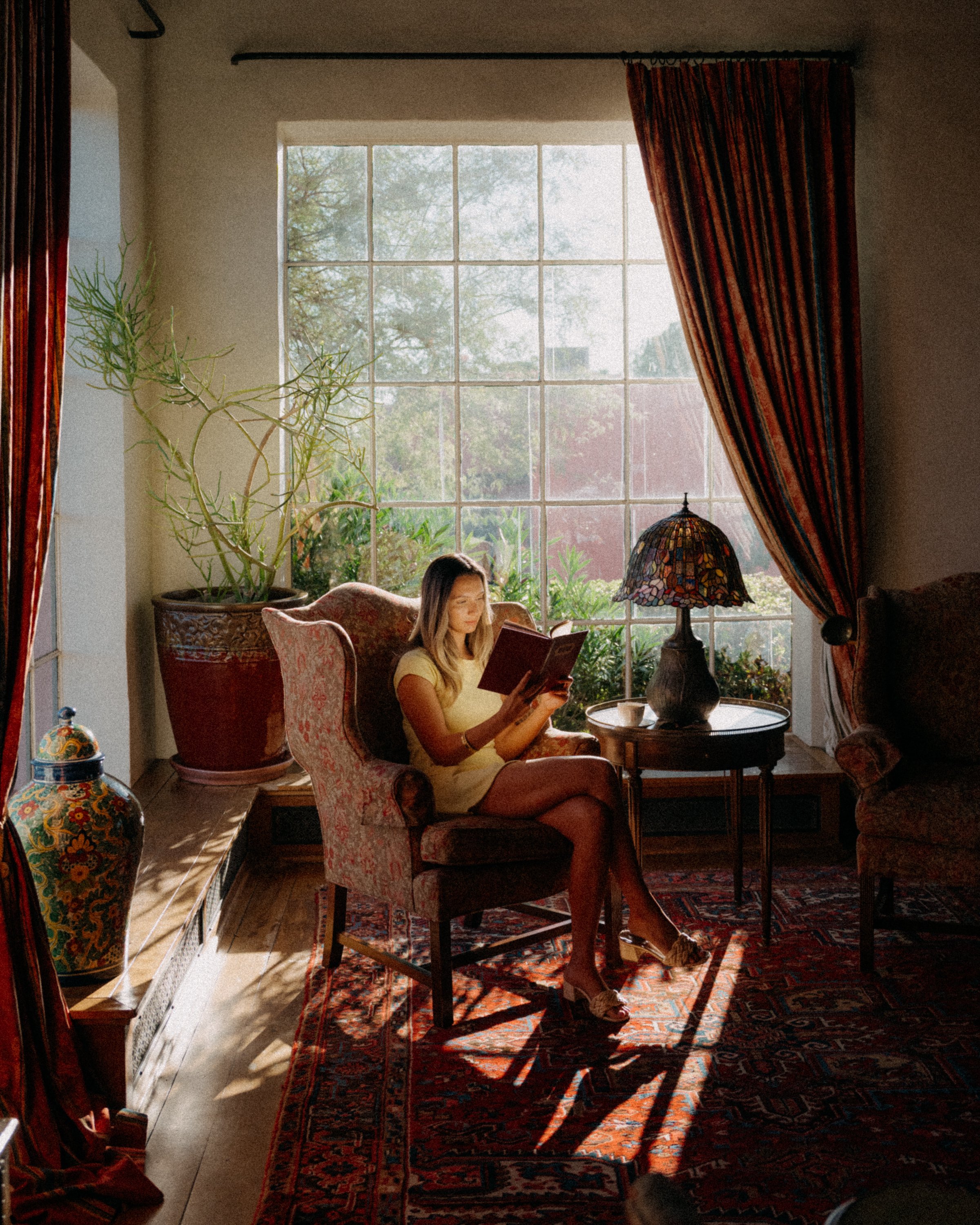A woman sitting in a patterned armchair reading a book in a cozy living room with large window, sunlight, plants, and a table lamp with stained glass shade.