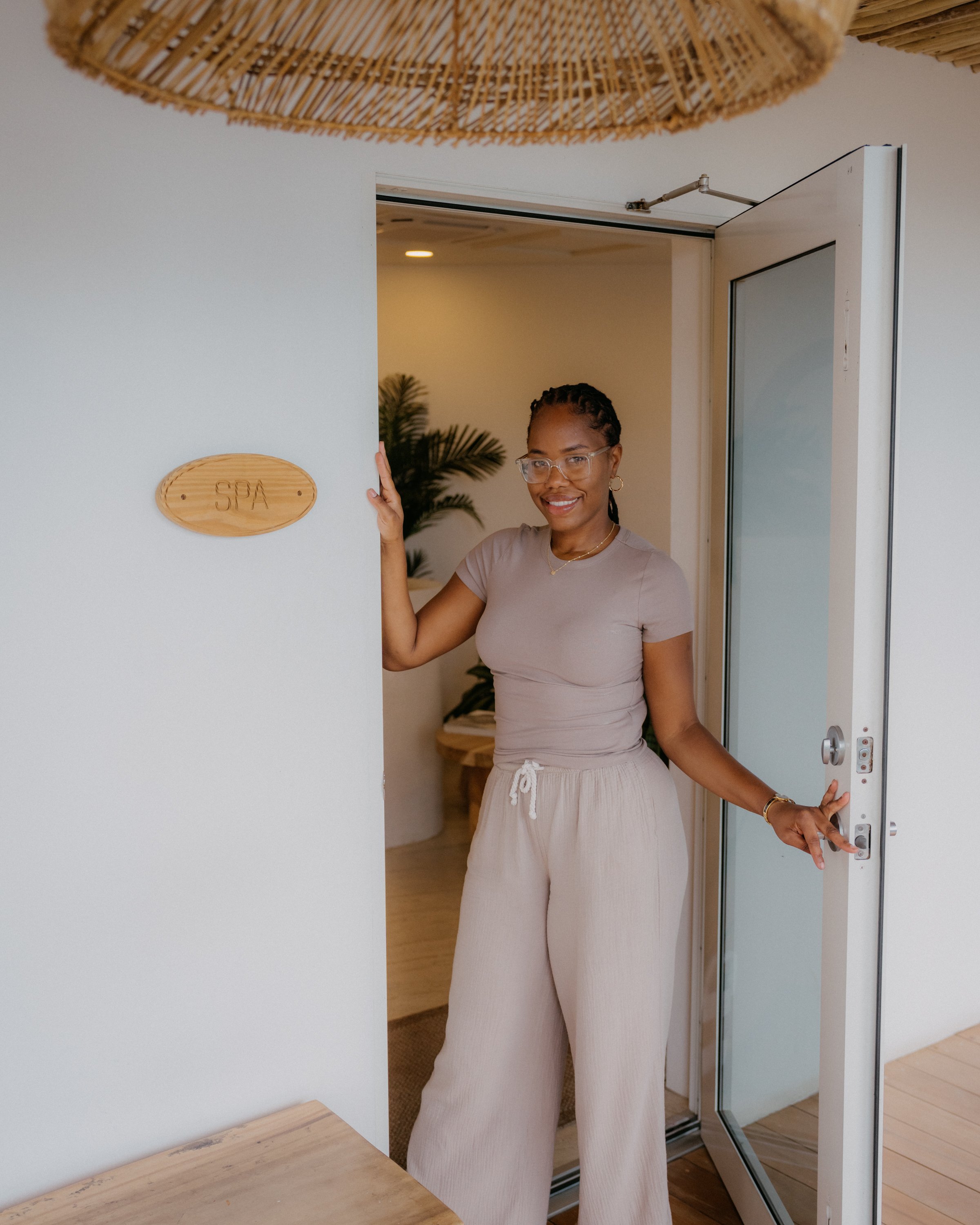 Smiling woman with glasses and earrings standing in a doorway, wearing beige loungewear, inside a wellness or spa setting.