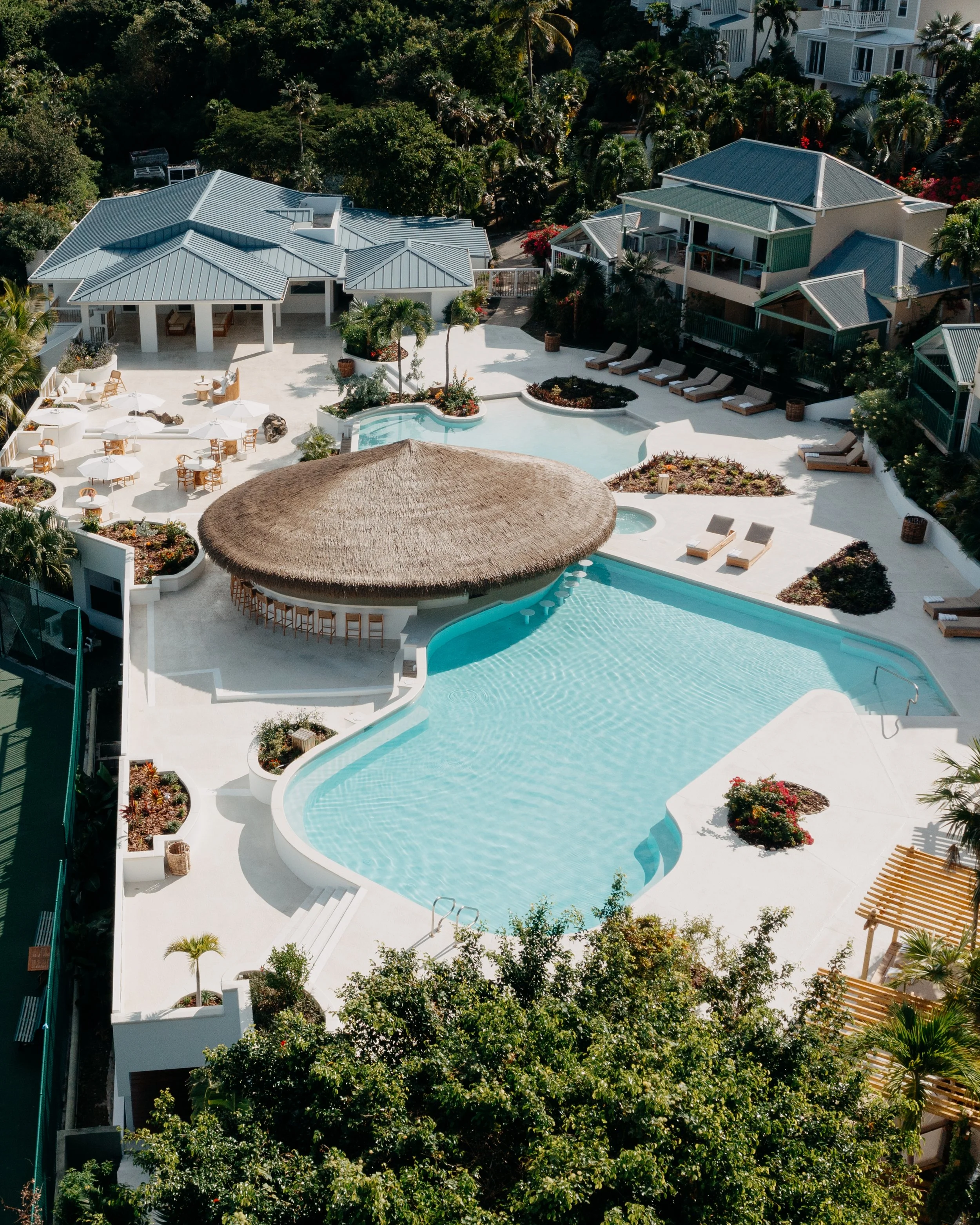 Aerial view of a luxurious pool area with lounge chairs, umbrellas, lush greenery, and multiple buildings with metal roofs surrounded by trees.