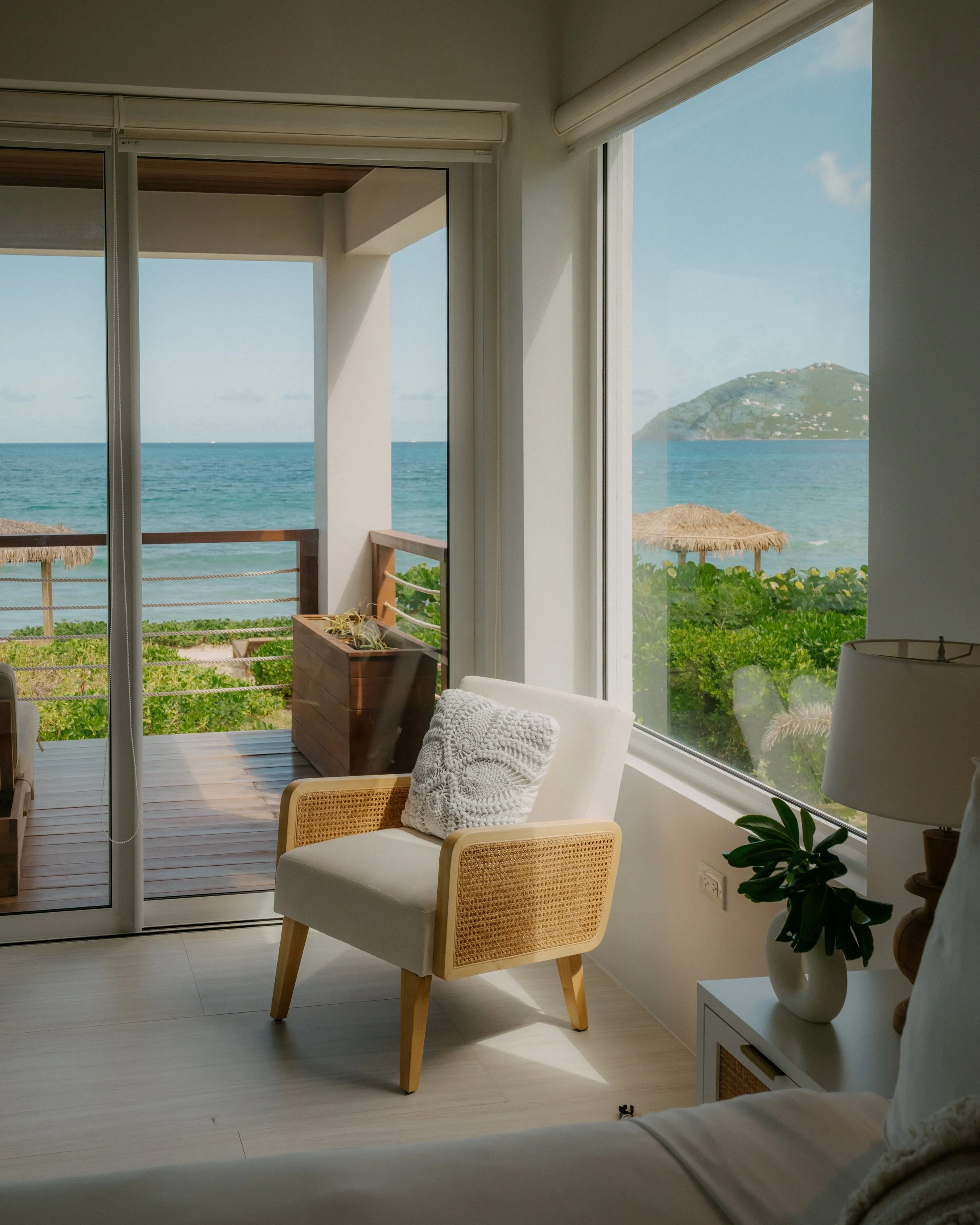 A cozy indoor space with large windows overlooking an ocean view, with a chair and a side table with a potted plant inside.