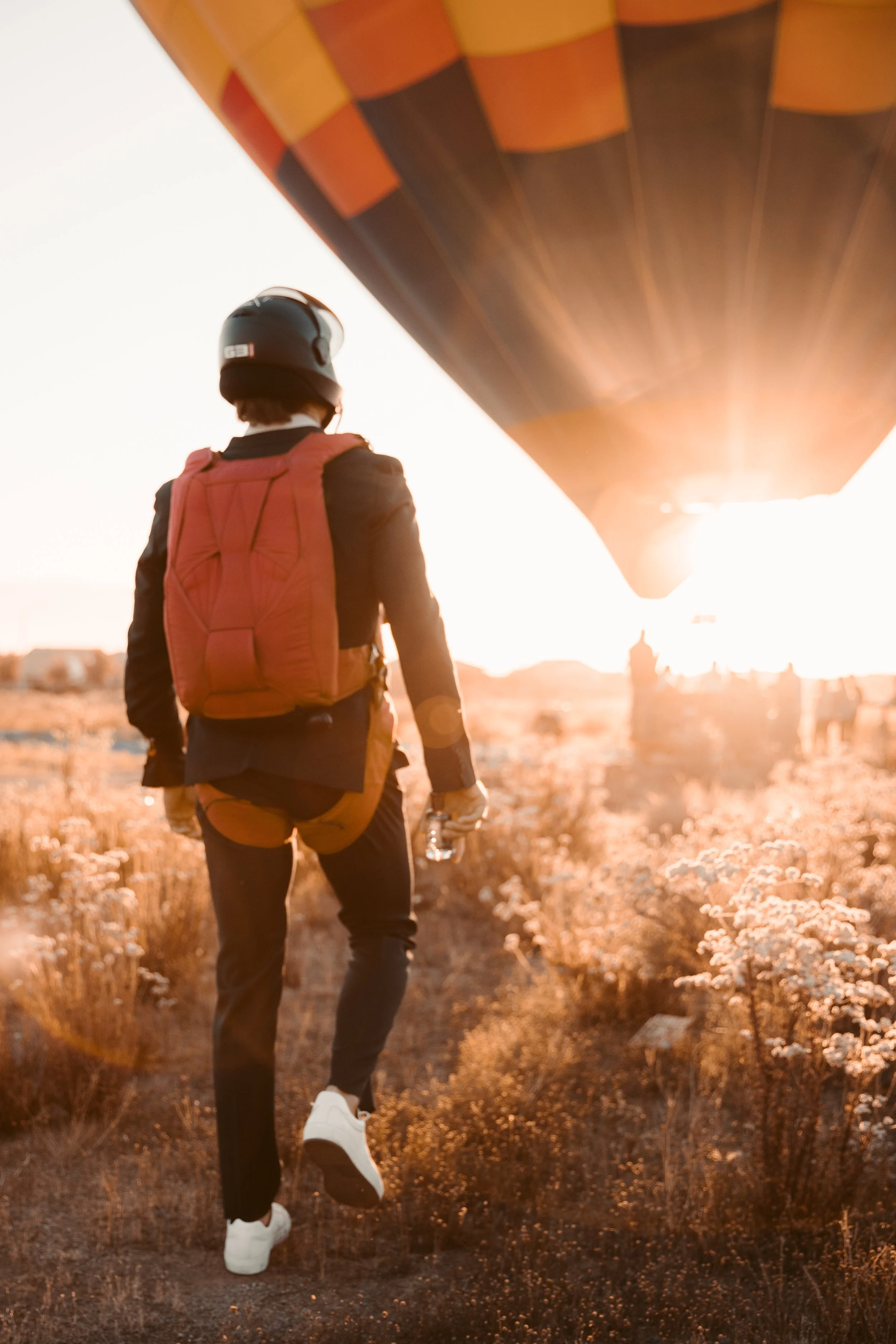 A person wearing a black helmet and red backpack walking through a field of wildflowers during sunset, with the hot air balloon in the sky.