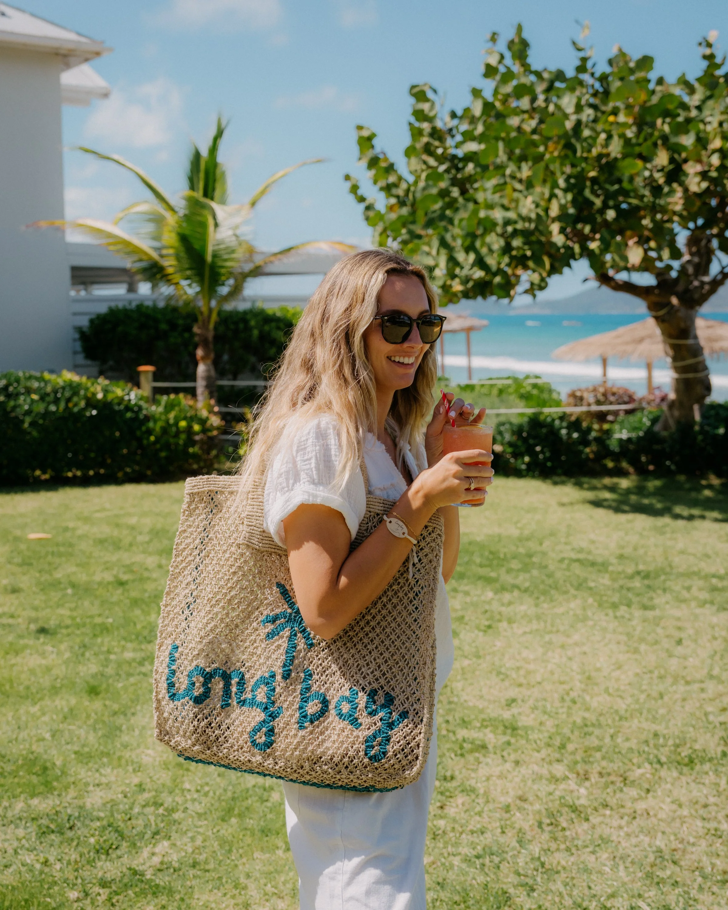 A woman wearing sunglasses and a white outfit is smiling while holding a drink. She has long blonde hair and is carrying a woven bag with blue embroidery. The background features a grassy area, trees, a white building, parasols, and the ocean under a