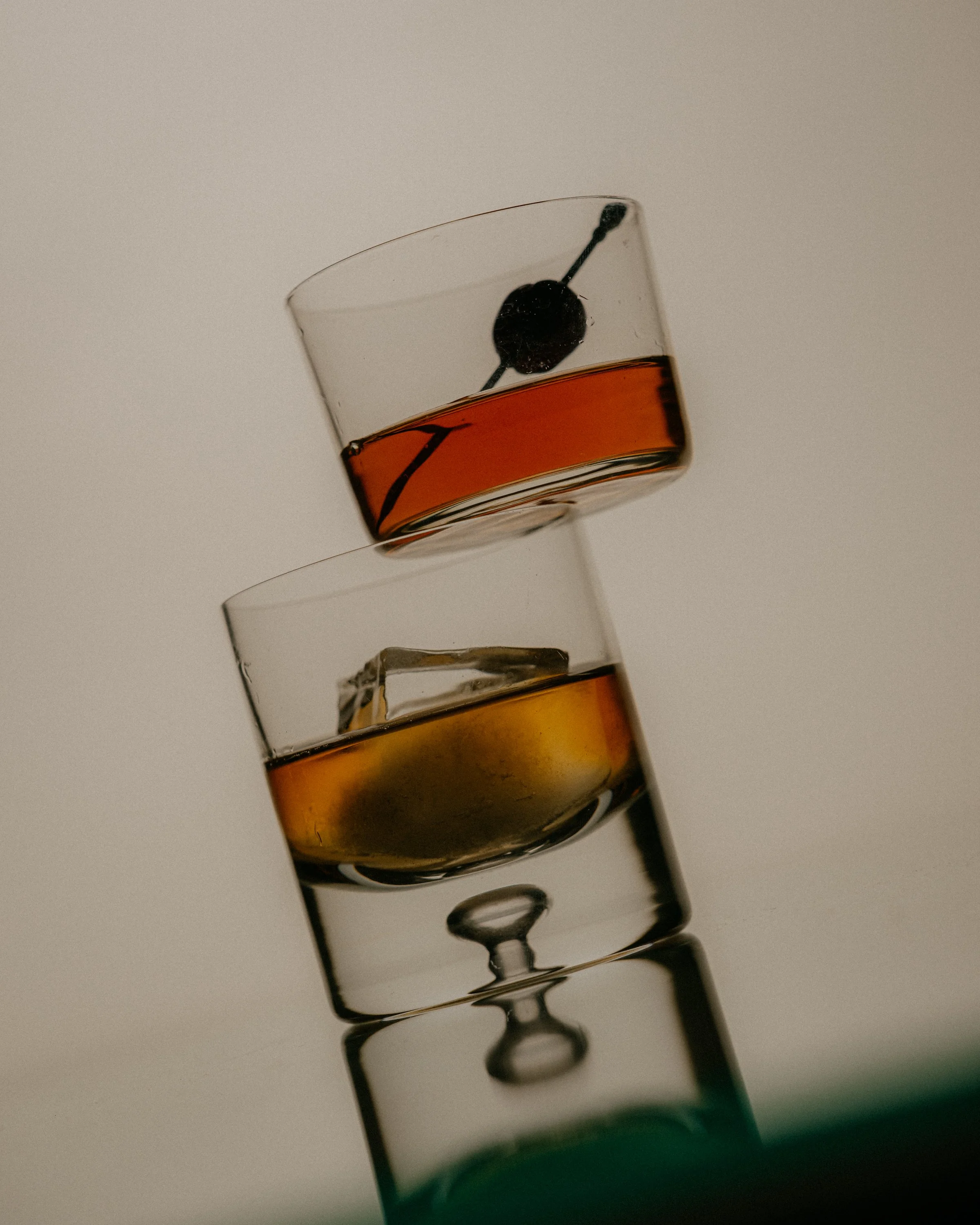 Close-up of a layered shot glass with amber-colored liquid and a black cherry garnishing the top. The shot glass is set against a plain, light background.