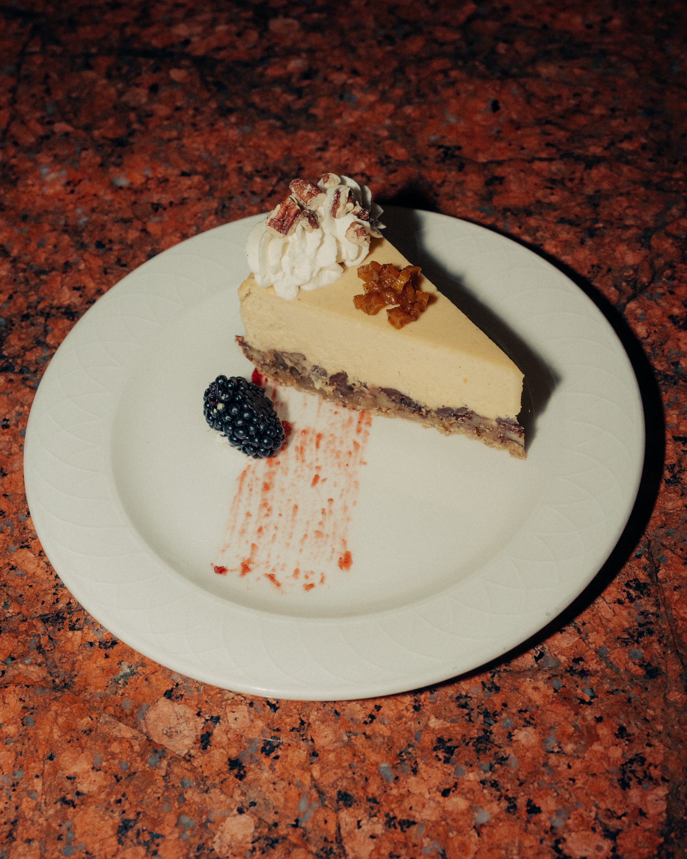 A slice of cheesecake with whipped cream and pecans on top, garnished with a blackberry and a drizzle of raspberry sauce on a white plate, placed on a red granite countertop.
