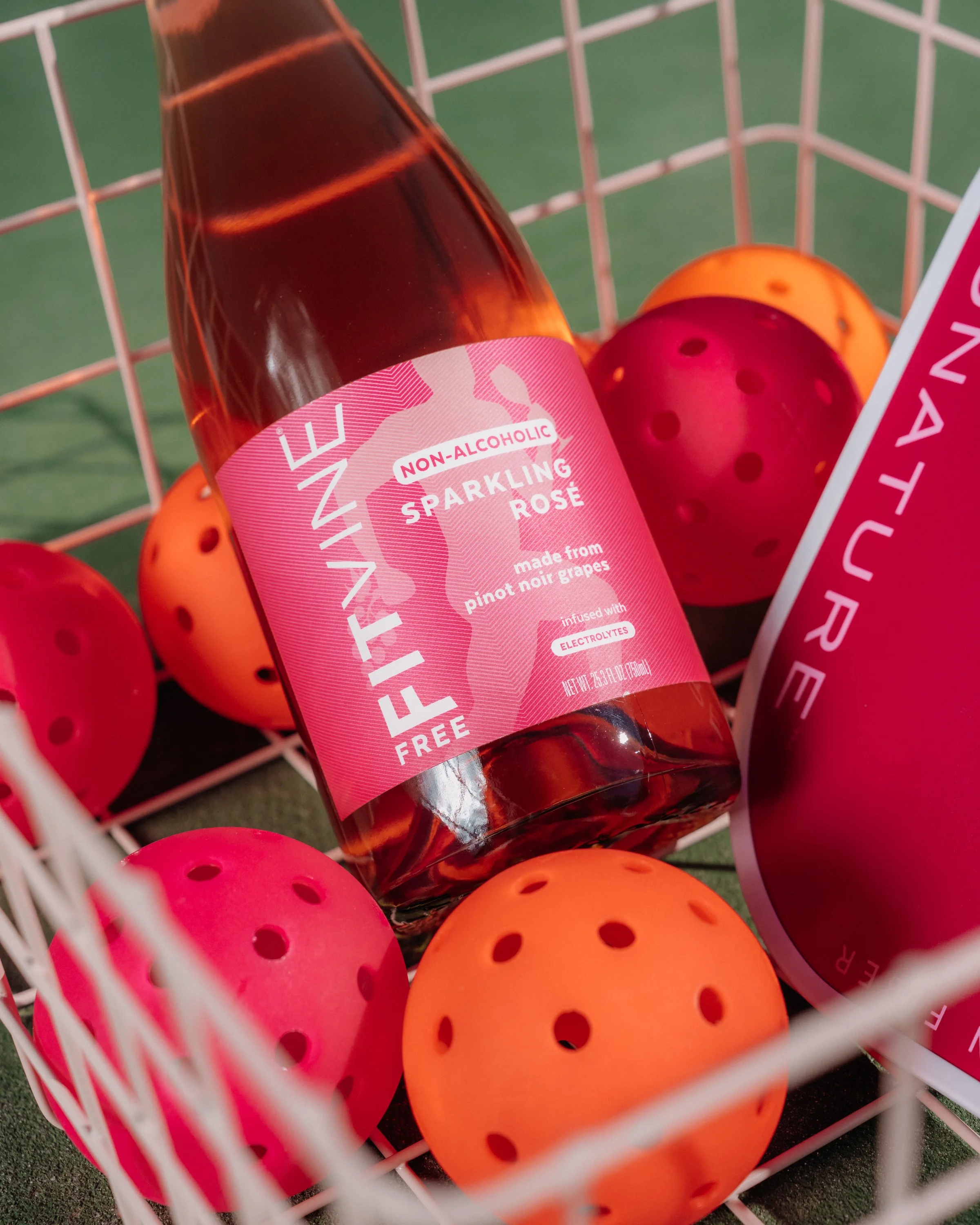 A bottle of non-alcoholic sparkling rosé wine in a pink label with a geometric pattern, surrounded by orange and pink tennis balls in a white basket.