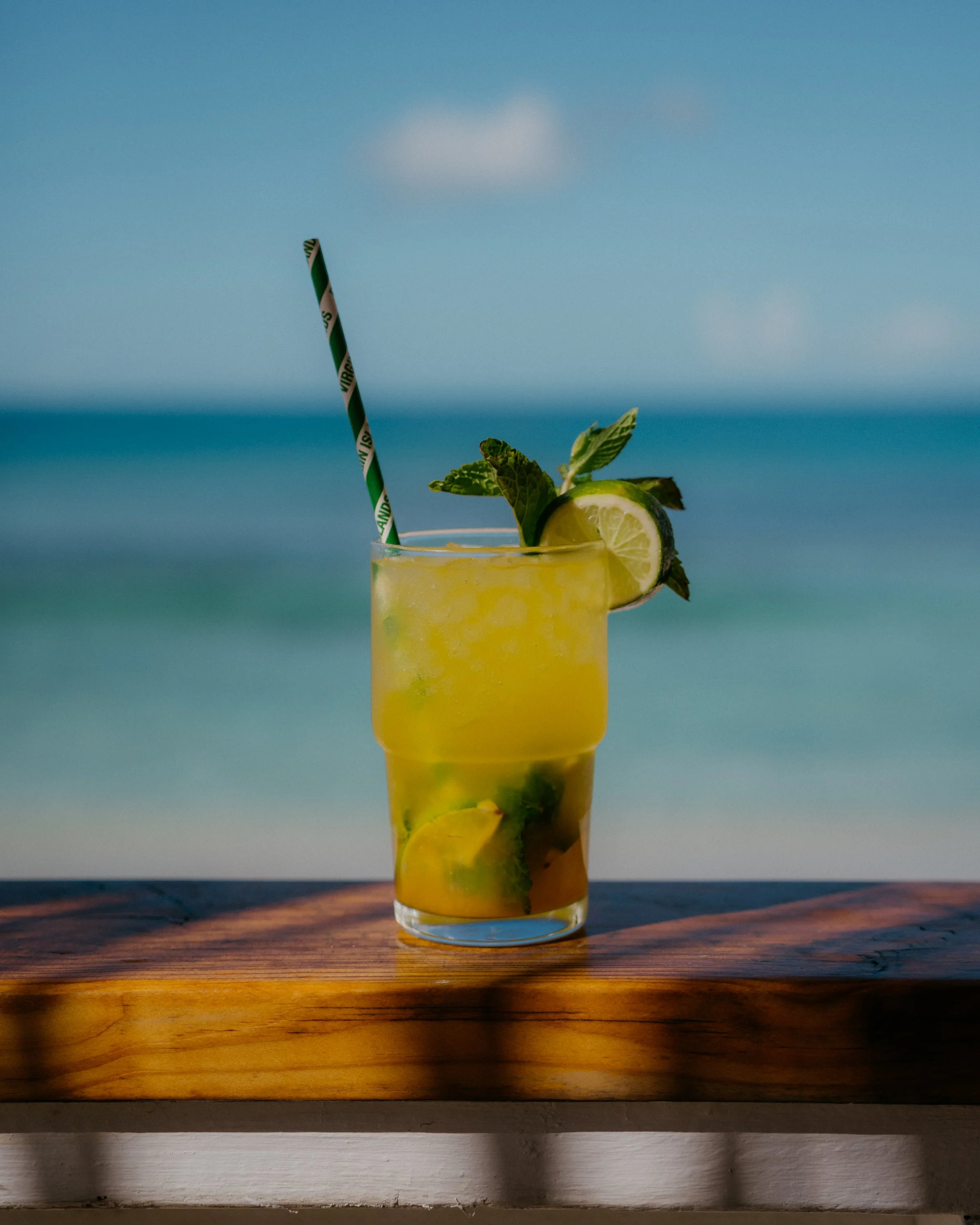 A tall glass of yellow lemonade garnished with lime slices, mint leaves, a green and white striped straw, and a lemon wedge on the rim, placed on a wooden surface with a beach and ocean in the background.