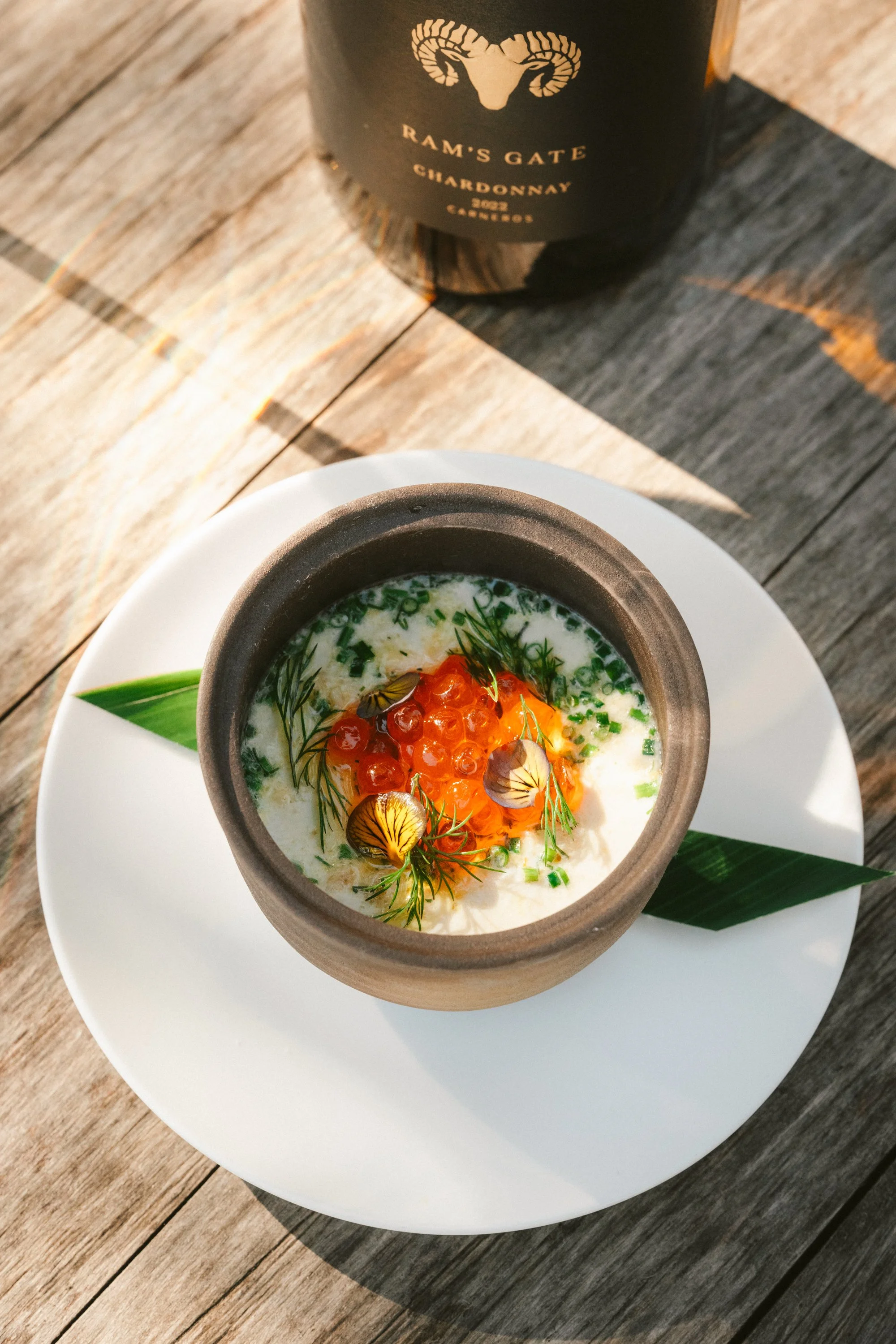 A bowl of savory dish topped with orange fish roe, garnished with dill and edible flowers, placed on a white plate with a green leaf. Behind, a bottle of Ram's Gate Chardonnay.