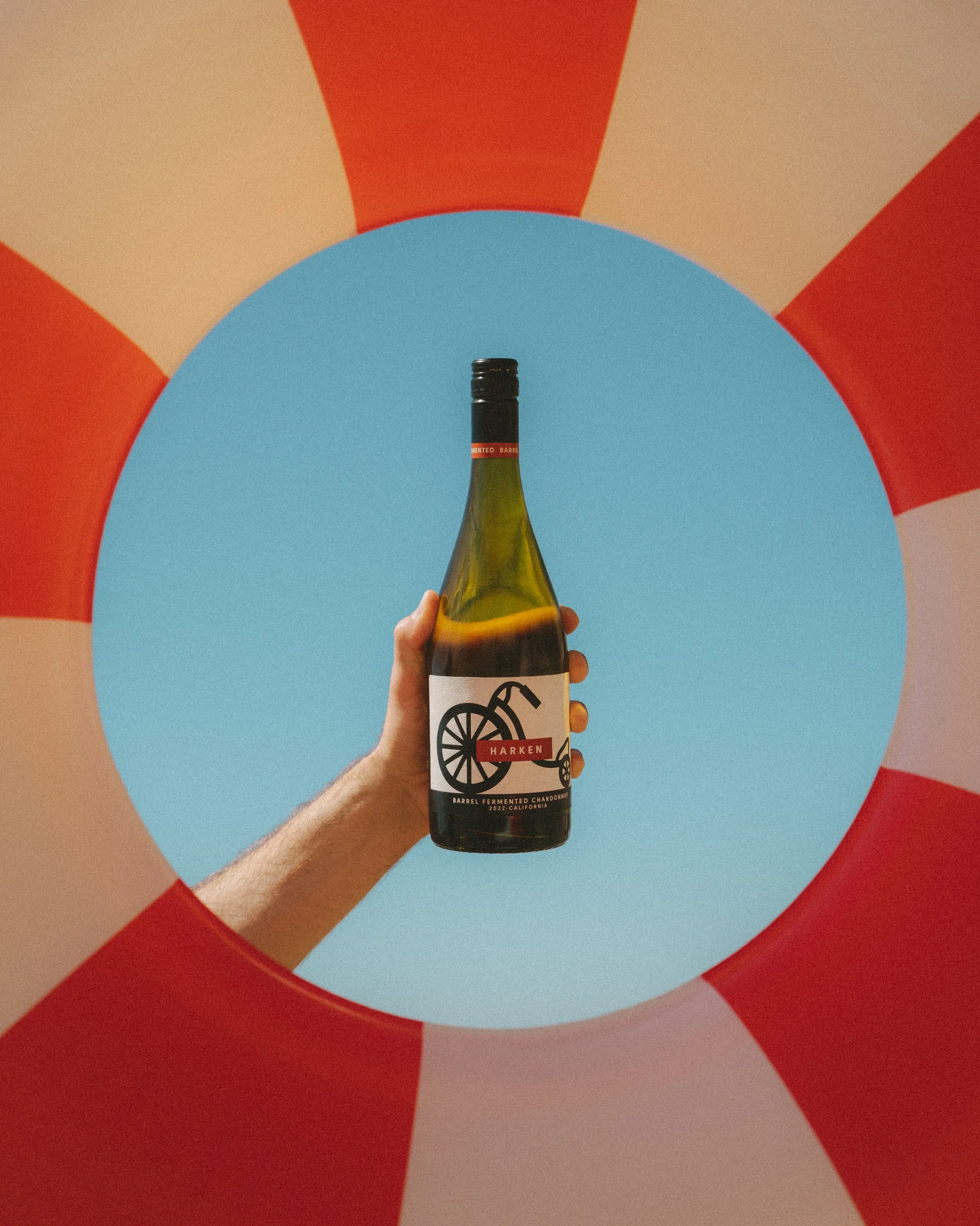A hand holding a bottle of Harken Barrel Fermented Chardonnay in front of a colorful geometric background with blue, orange, red, and beige shapes.