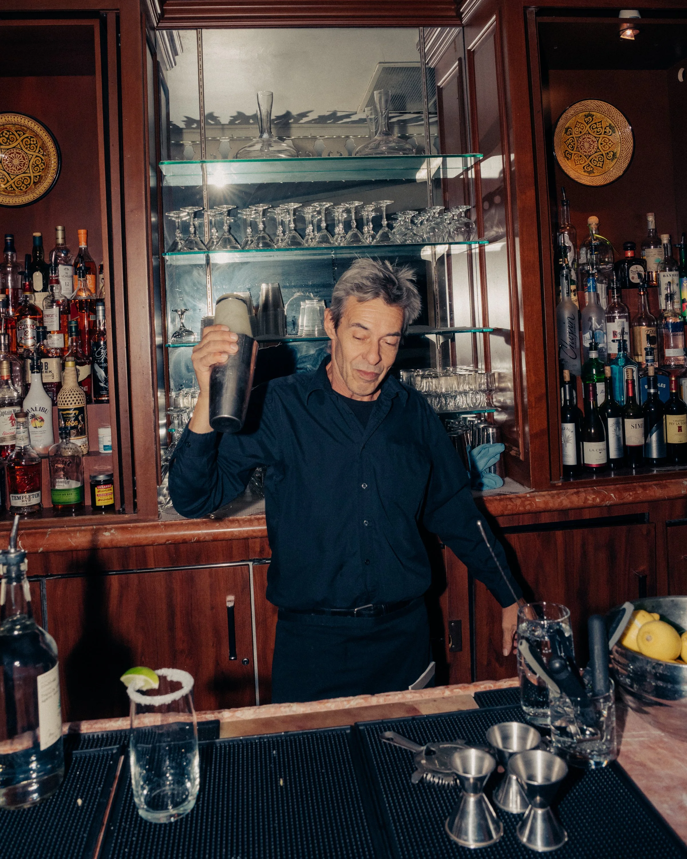 A man with gray hair in a black shirt working behind a bar, shaking a cocktail shaker. The bar has wooden cabinetry, shelves with liquor bottles, and glassware including wine glasses and cups. There are bar tools and a glass of water with a lime on t