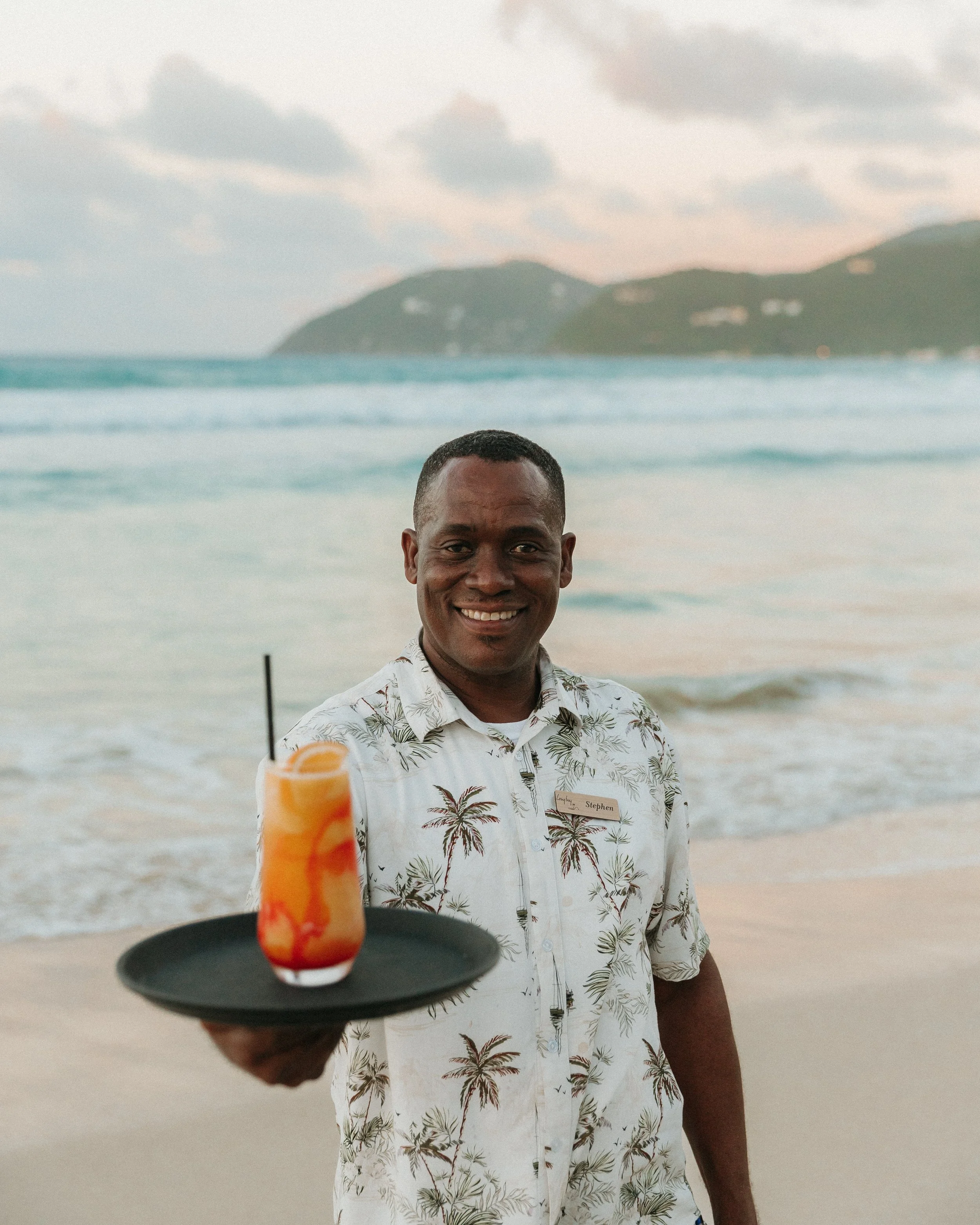 A man with short hair wearing a white Hawaiian shirt featuring palm trees, standing on a beach with the ocean and hills in the background. Someone is holding a tray with a cocktail topped with an orange slice in the foreground.