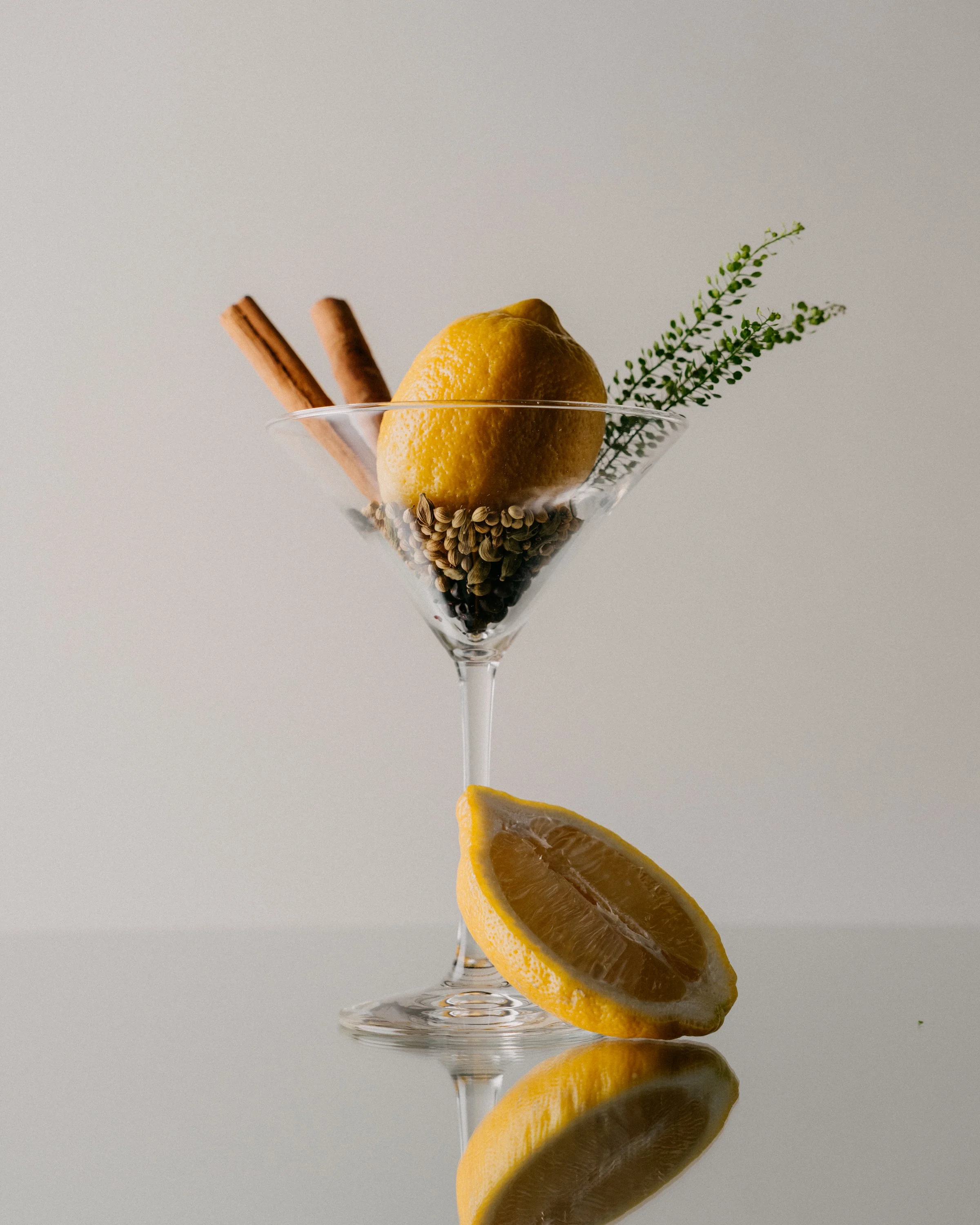 A martini glass filled with black peppercorns, garnished with a lemon wedge, a sprig of green herbs, and two cinnamon sticks, set against a plain background.