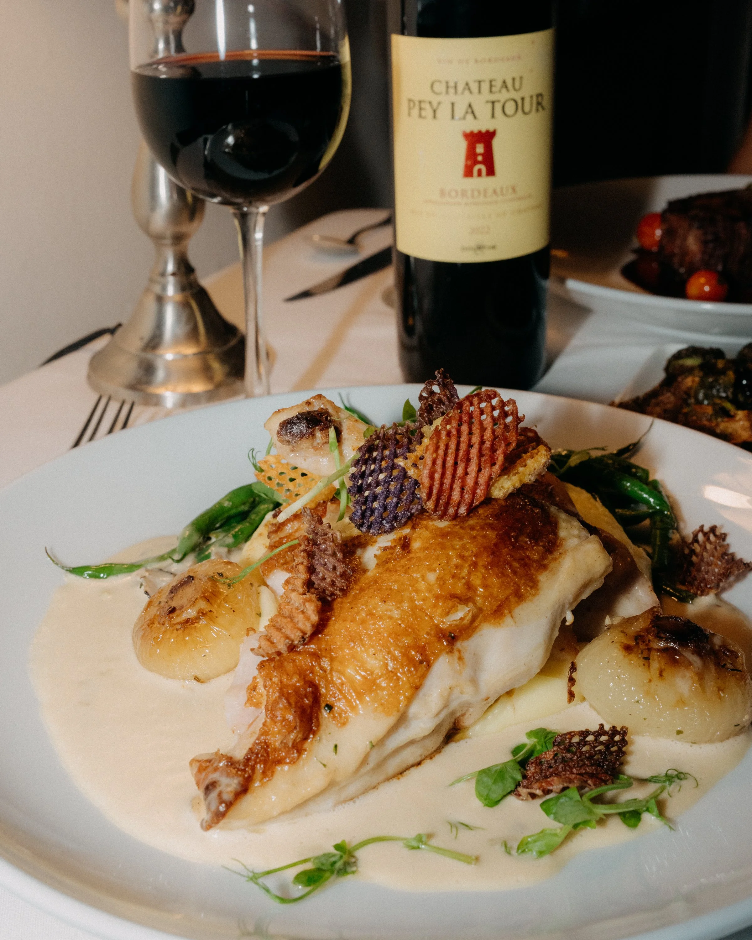 A close-up of a gourmet dish with a piece of fish topped with crispy garnish, served with green vegetables and potatoes. In the background, a glass of red wine, a bottle of Château Pey La Tour Bordeaux wine, and a plate of roasted meat with cherry to