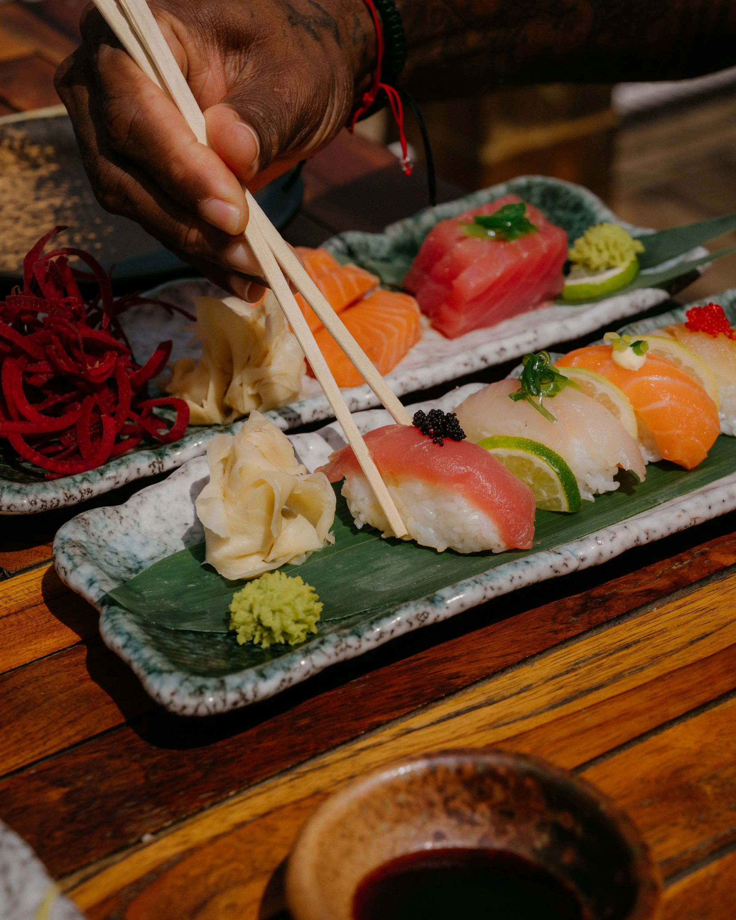 A hand using chopsticks to pick up sushi pieces on a ceramic plate with various types of sashimi, garnishes, and condiments, set on a wooden table.