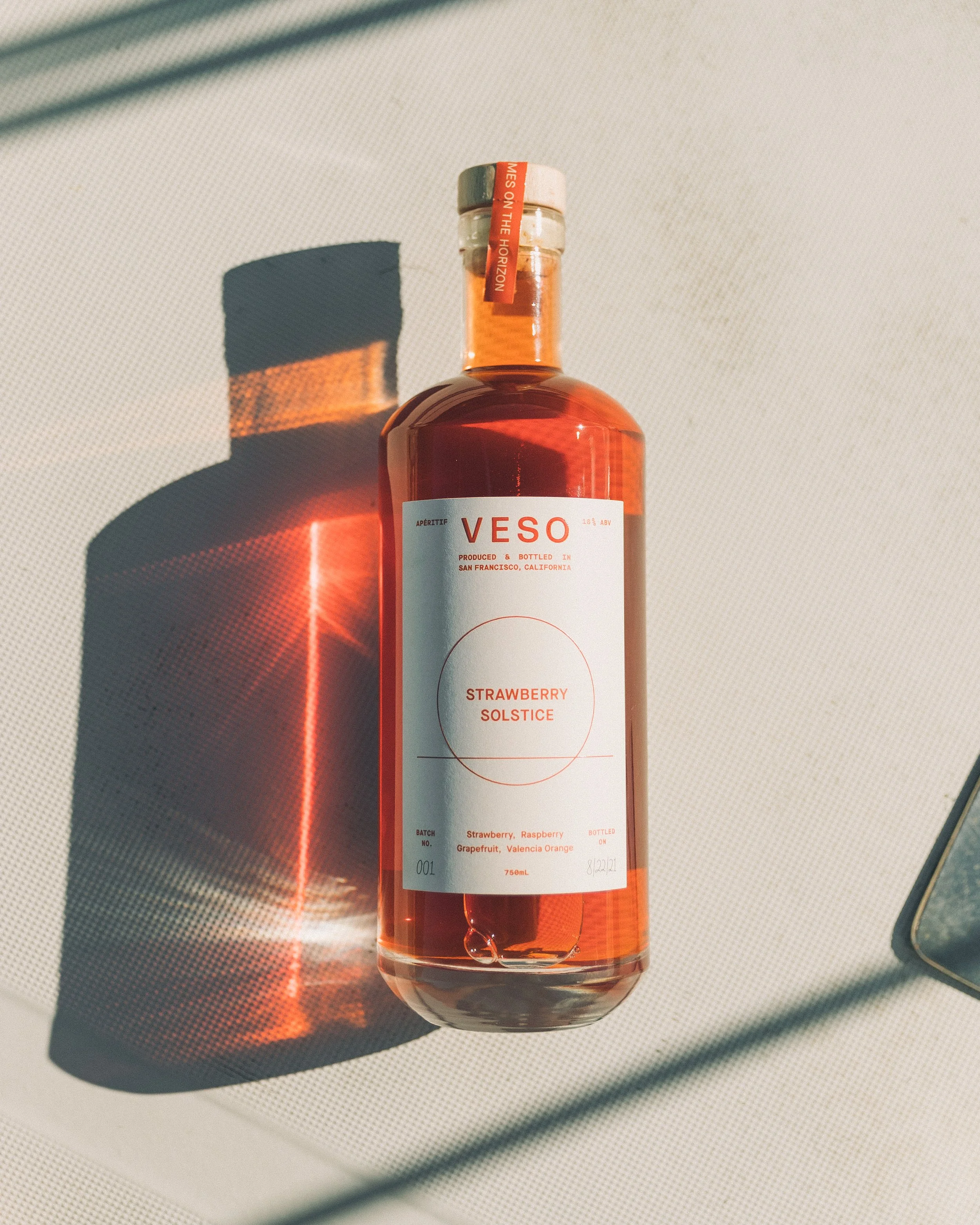 A bottle of strawberry solstice aperitif with a white label showing the flavor, alcohol content, and origin, placed on a white surface with a shadow cast to the left.