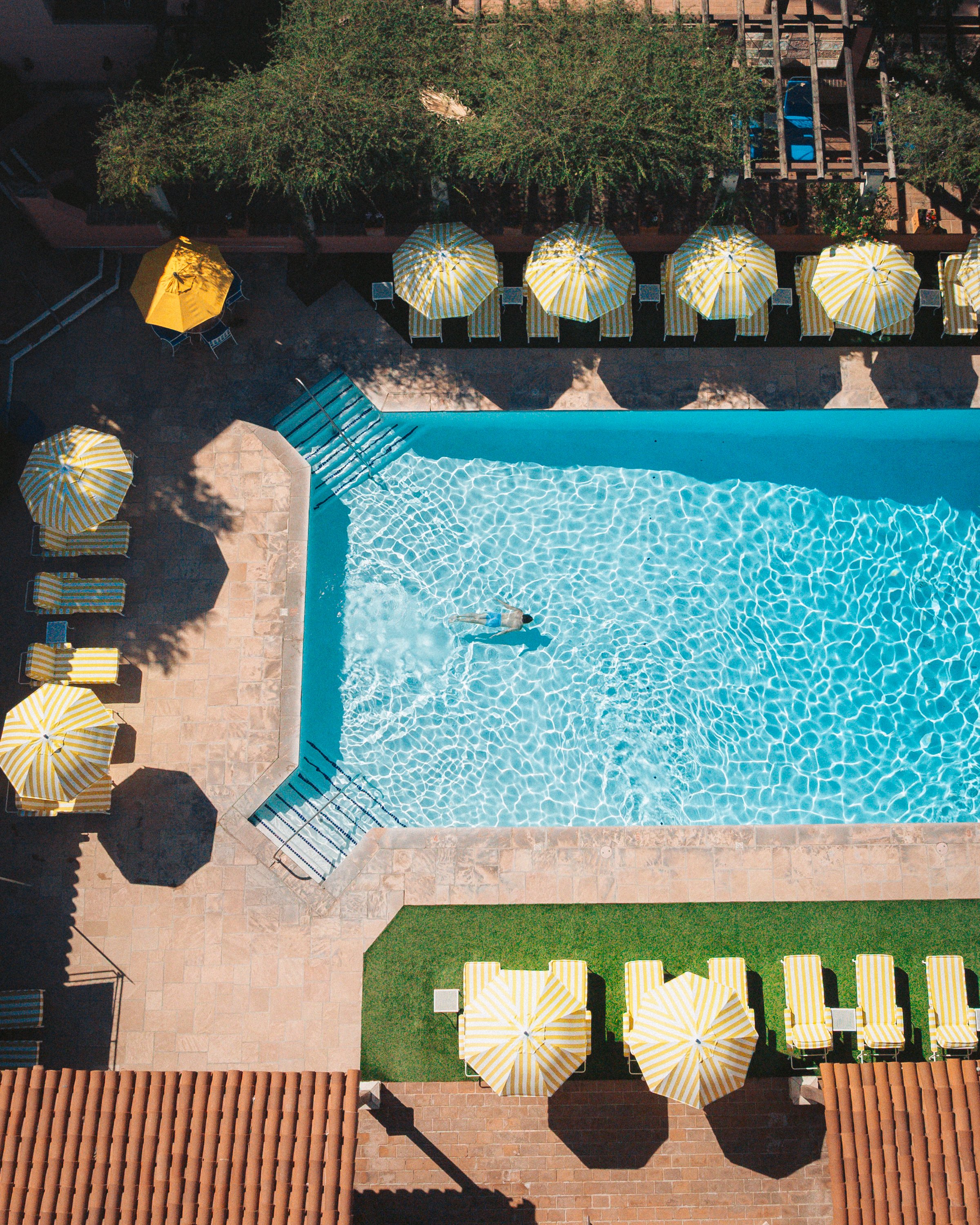 An aerial view of a swimming pool area with yellow and white striped umbrellas and lounge chairs on tiled and artificial grass surfaces, with trees and a wooden deck in the background