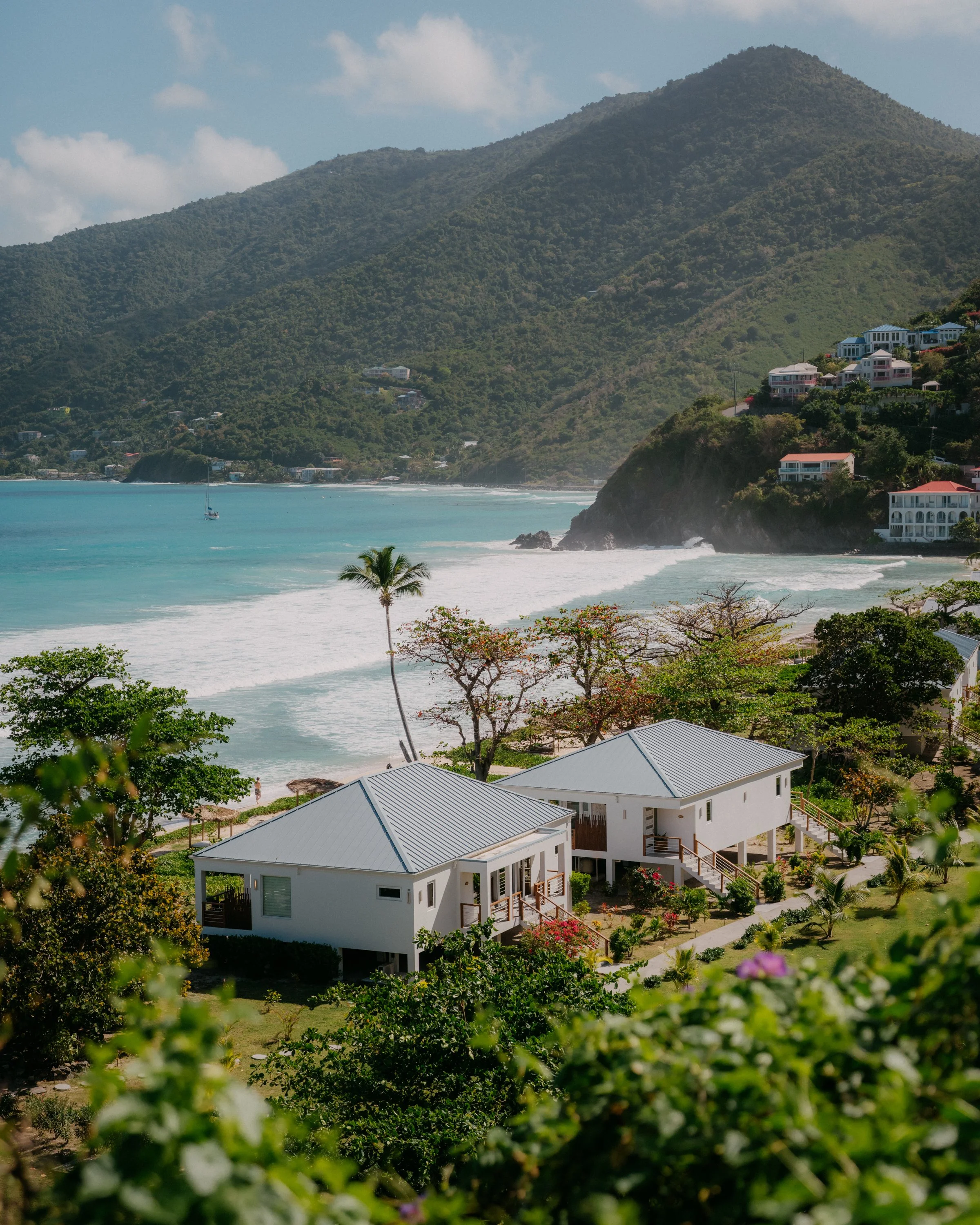 A coastal scene with white houses on a hillside, lush greenery, a sandy beach, turquoise sea, and a mountain in the background under a partly cloudy sky.