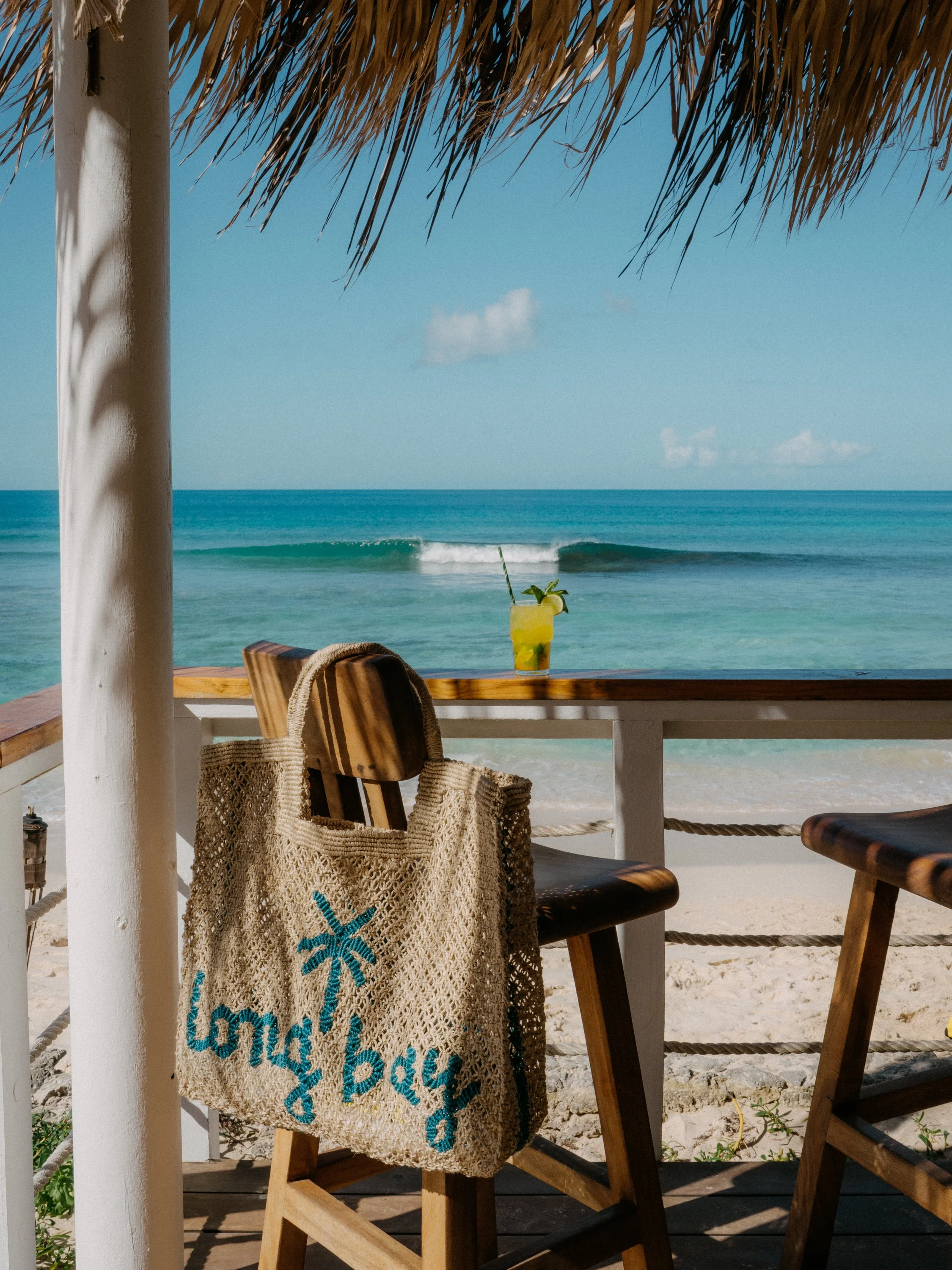 Beachside bar with wooden chairs, a straw hut roof, and a view of the ocean with small waves and blue sky, a tall glass of yellow tropical drink with a lime wedge and mint leaves on a table.
