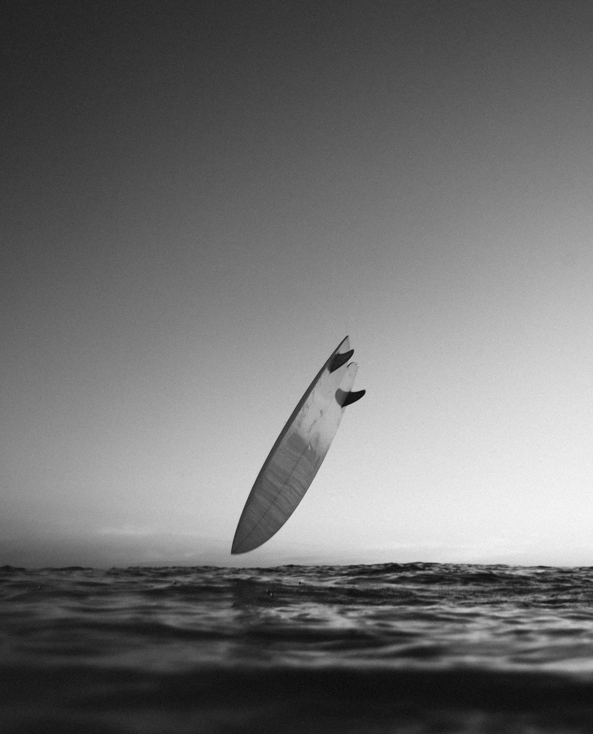 Black and white photo of a surfboard partially submerged in the ocean, with the sky above and the horizon in the distance.