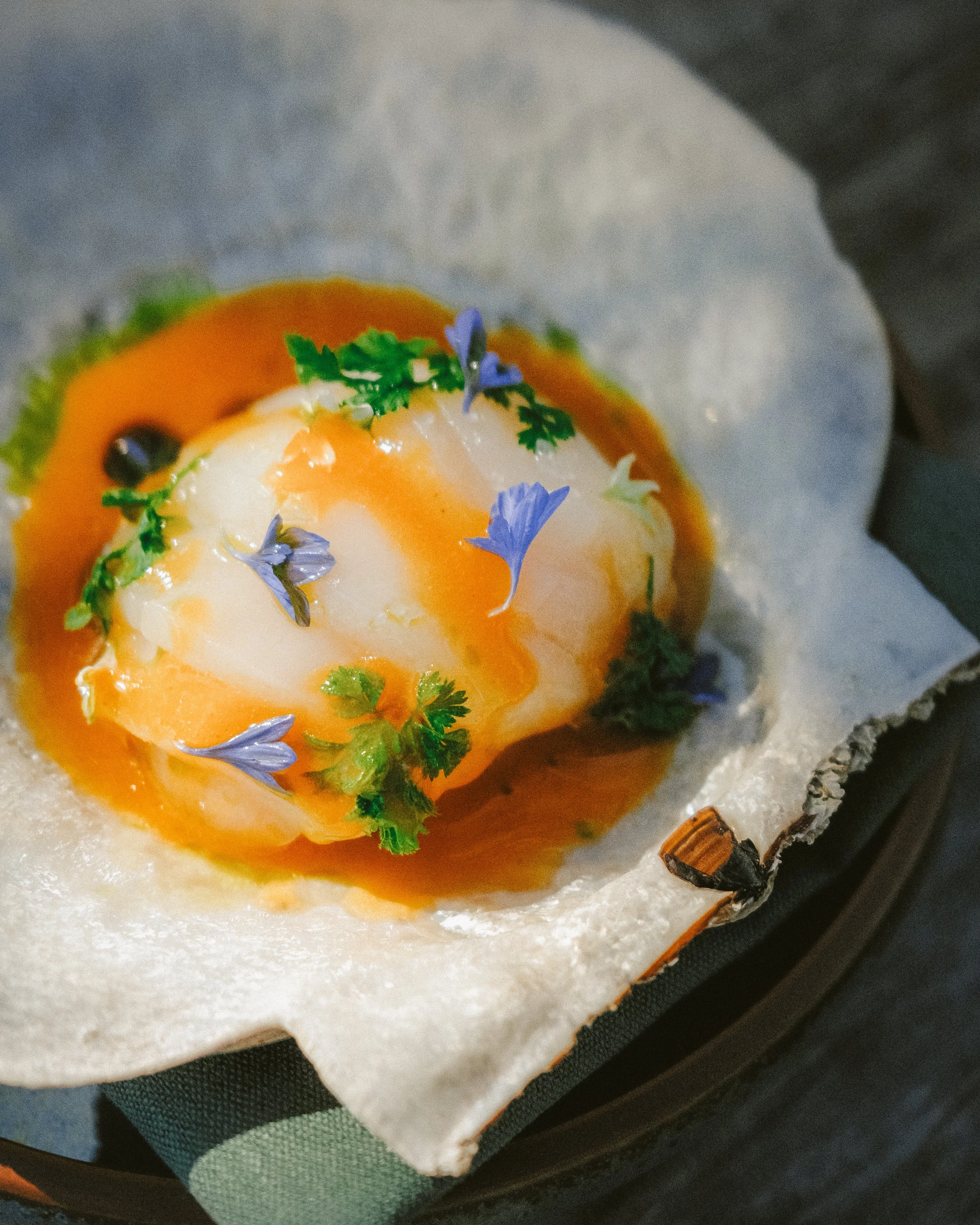 A soft white bun with a poached egg, topped with orange sauce, fresh herbs, and purple edible flowers, served on parchment paper on a black tray.