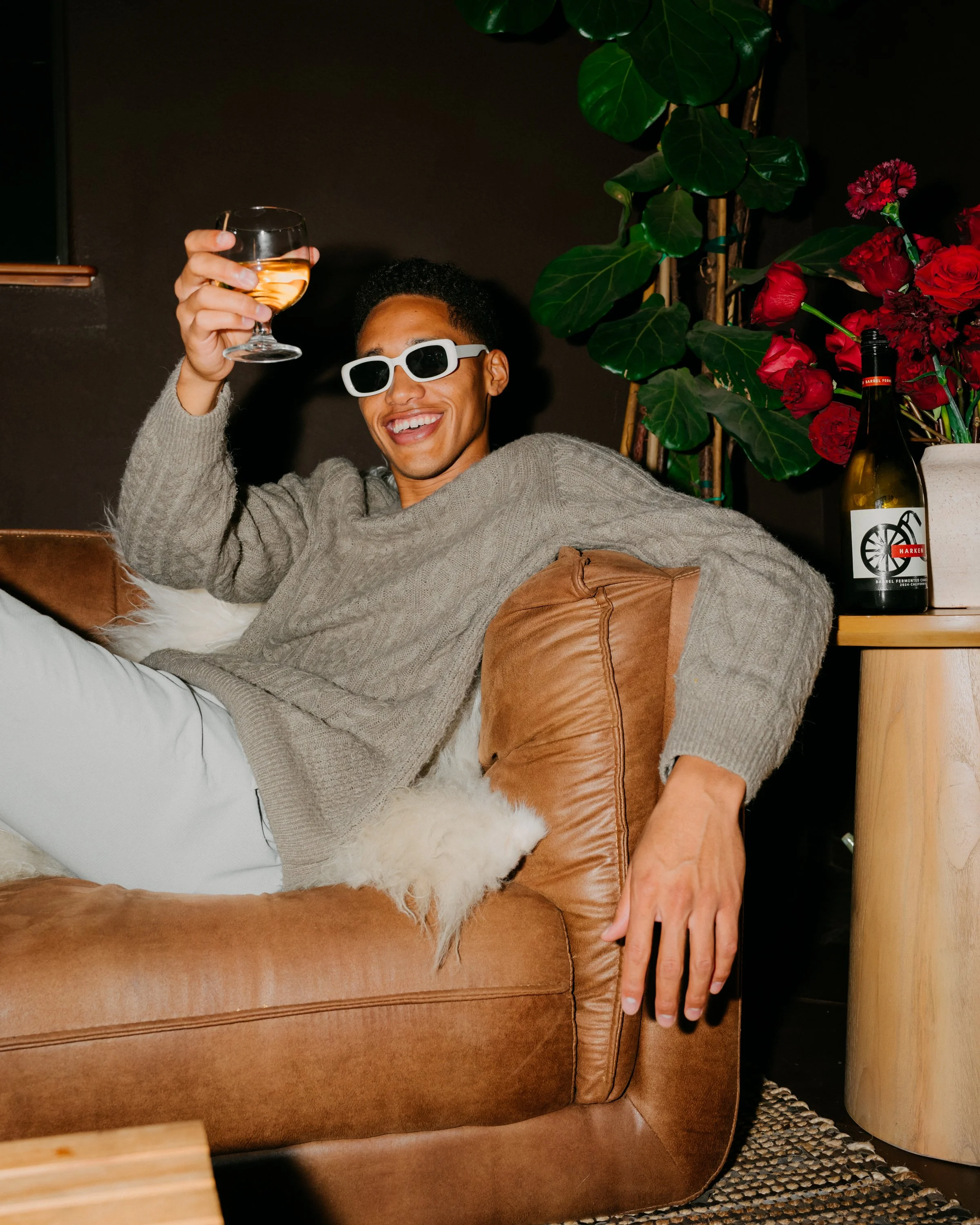 A person lounging on a brown leather sofa, smiling, wearing white sunglasses and a gray sweater, holding a glass of wine. In the background, there is a green plant, a bouquet of red flowers, and a bottle of wine or beer.