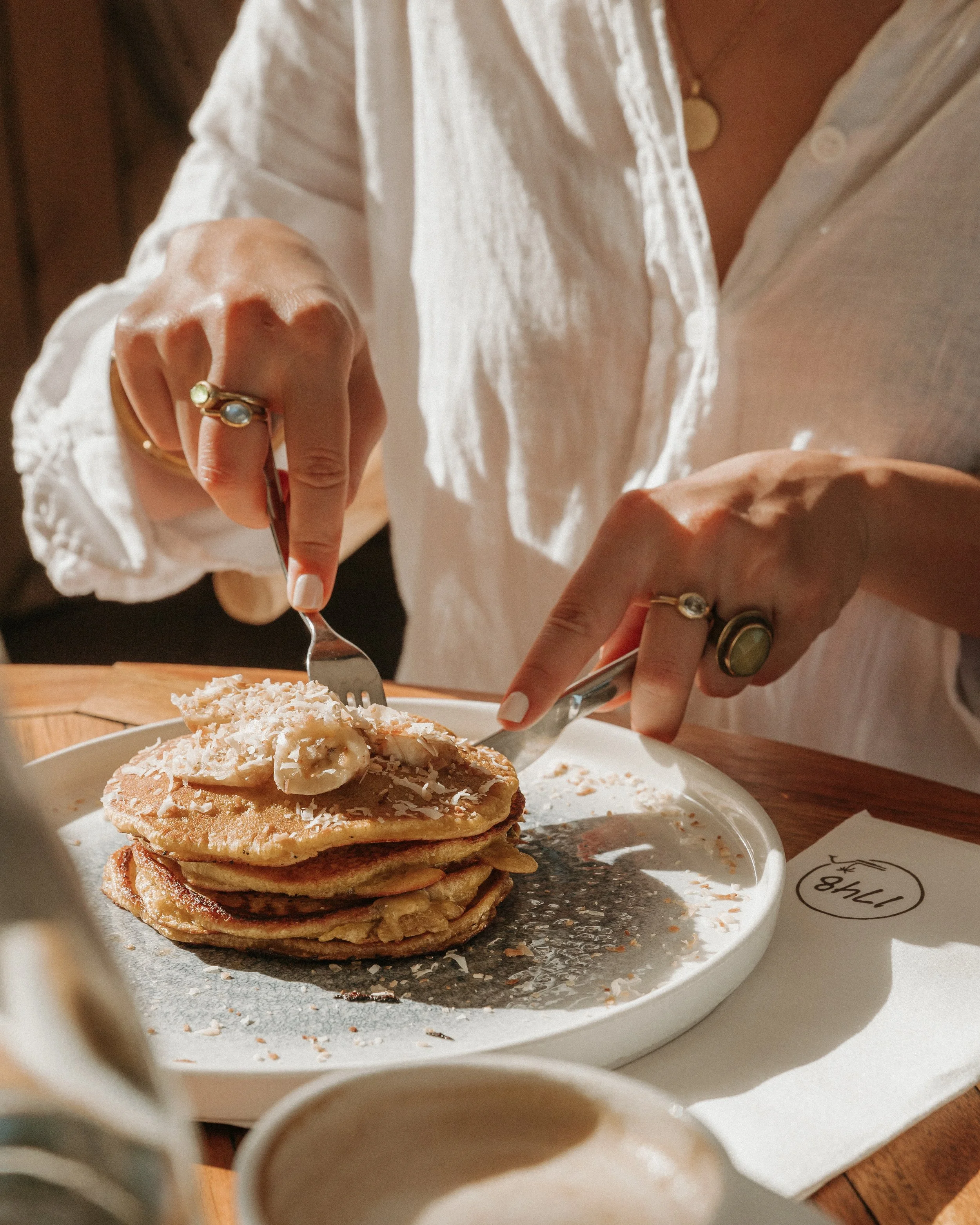 A person is serving a thick stack of pancakes topped with shredded coconut and a dollop of butter from a white dish. The person is wearing a white shirt with rolled-up sleeves and has rings on their fingers, while holding a fork and knife. A cup and 