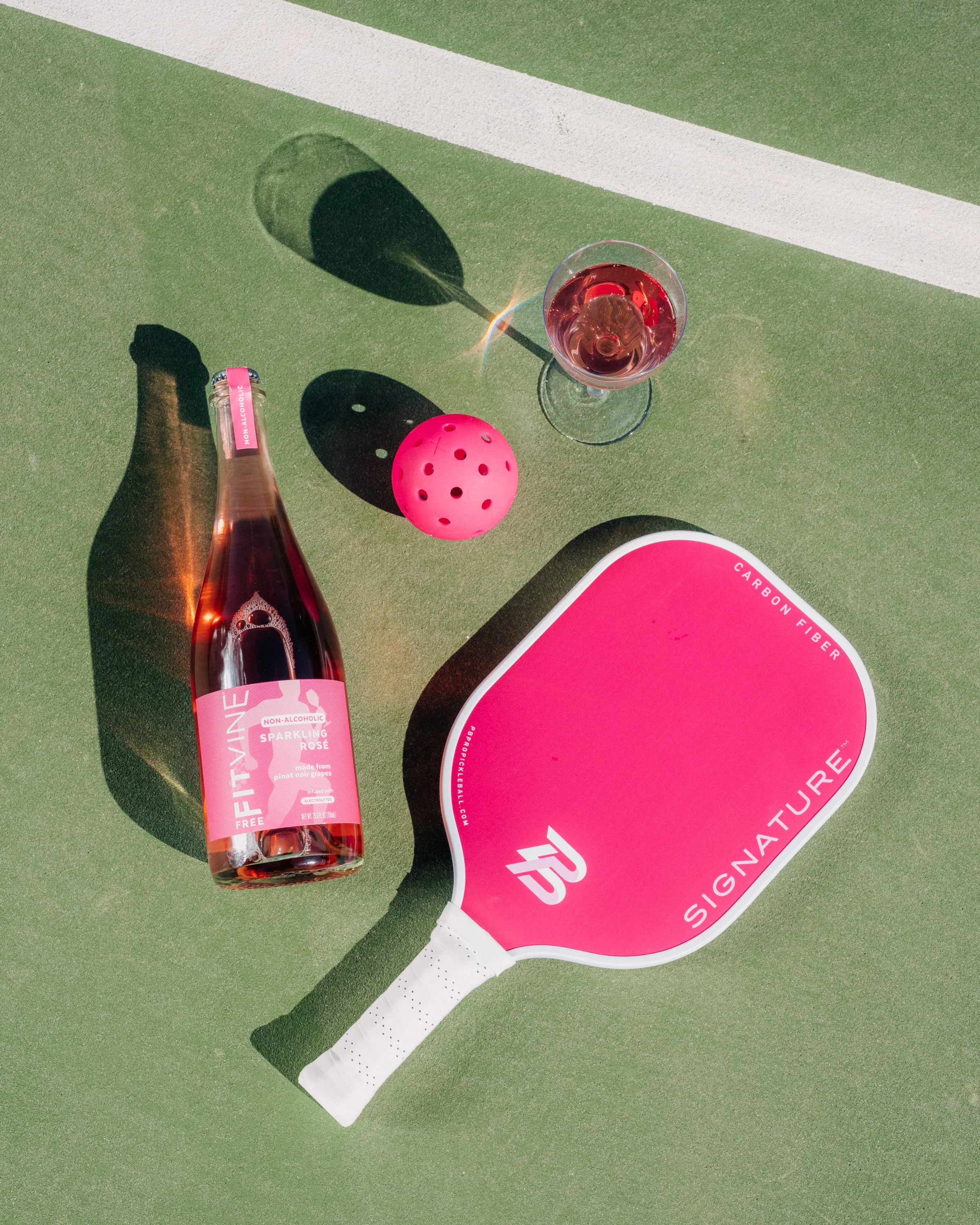A green tennis court surface with a pink non-alcoholic sparkling rosé wine bottle, a glass of rosé wine, a pink ball with holes, and a pink and white tennis racket with the words "SIGNATURE" on its strings.