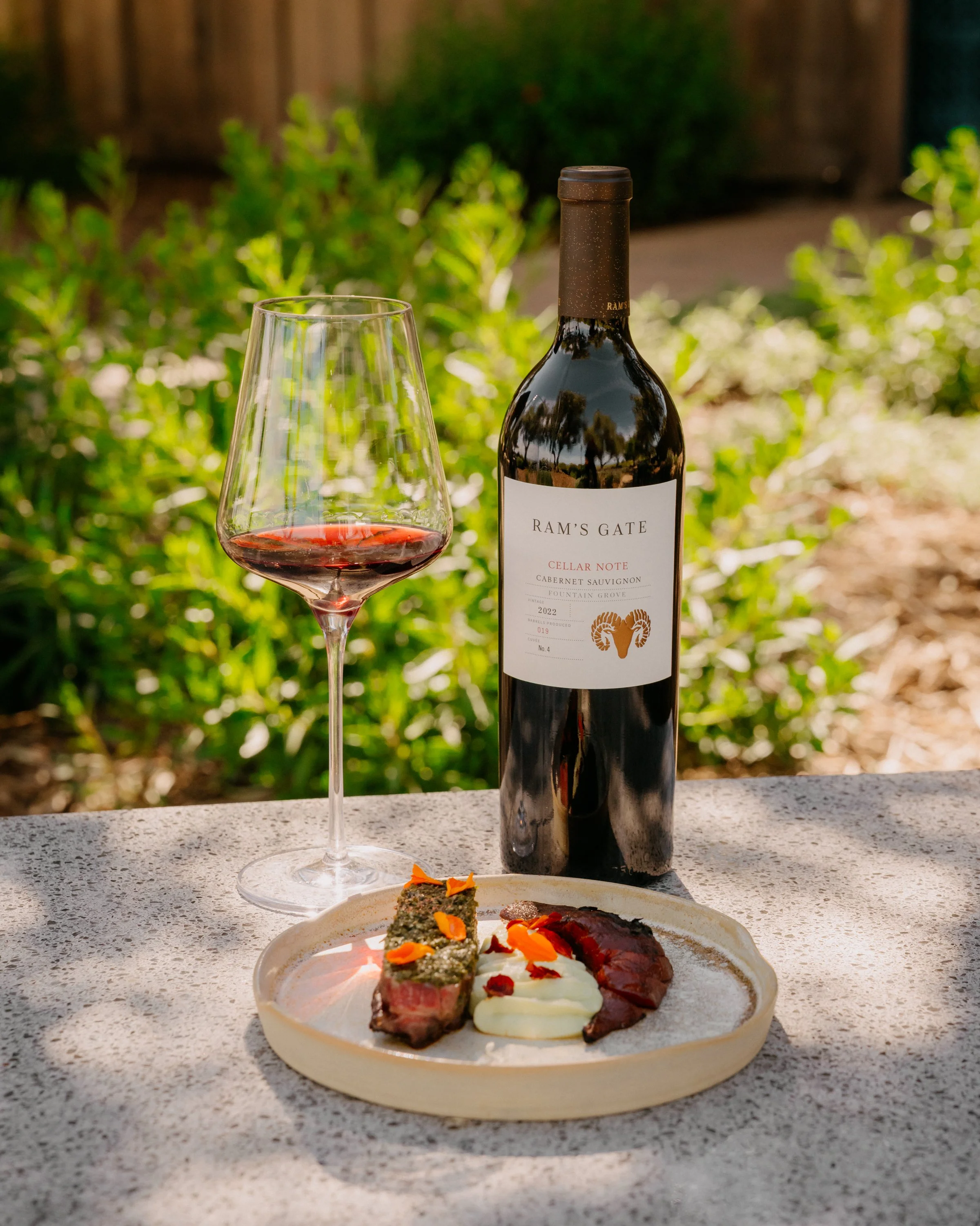 A glass of red wine, a bottle of Ram's Gate wine, and a plate of assorted meats and cheeses on a table outdoors, with greenery in the background.