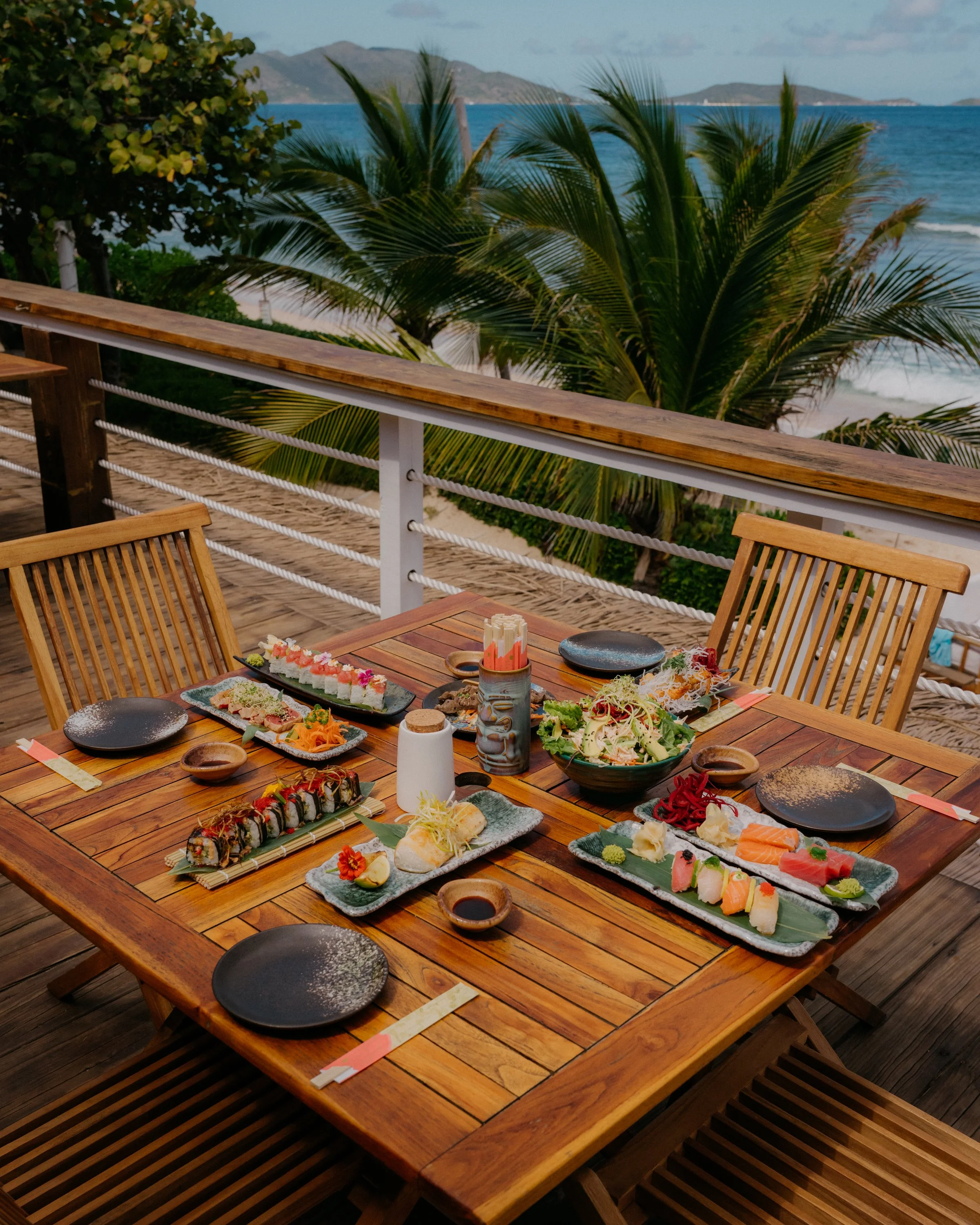 A wooden outdoor table filled with various plates of sushi, sashimi, and other Japanese dishes, set on a terrace overlooking palm trees, the ocean, and distant islands.