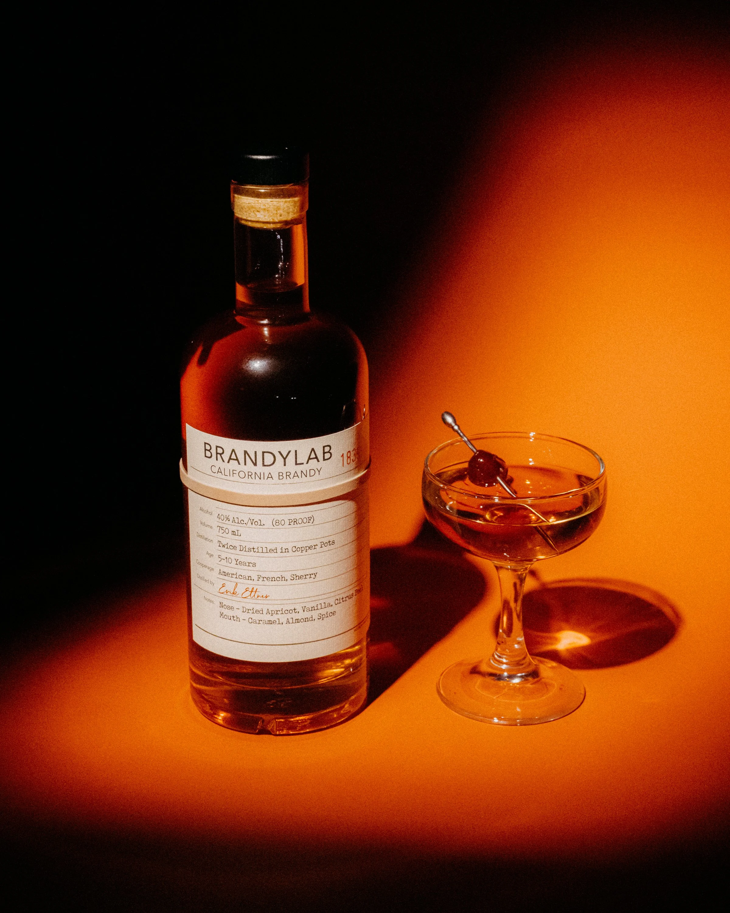 A bottle of Brandy labeled 'California Brandy' and a cocktail glass with a cherry on top, set against a dark background with warm orange lighting.