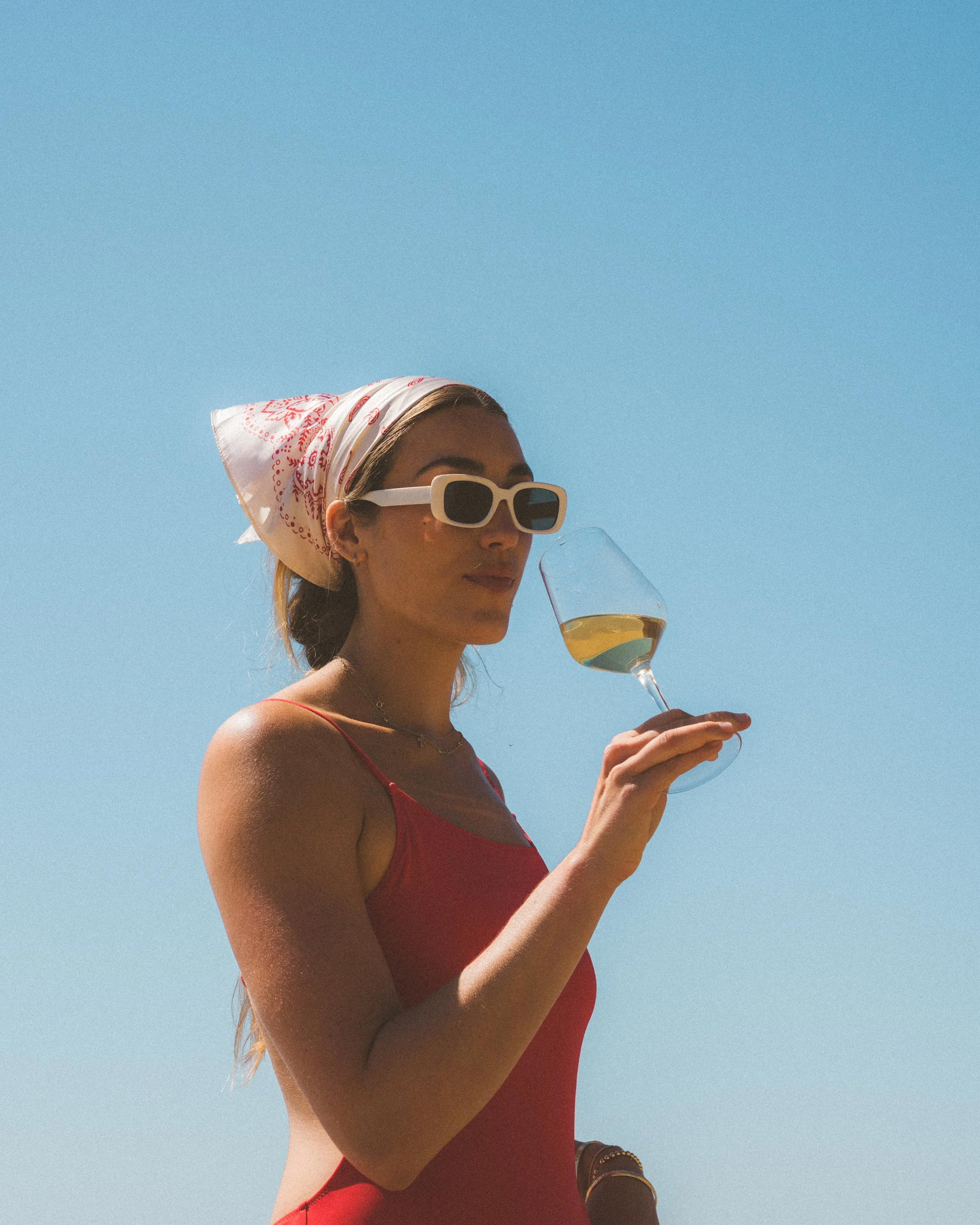 A woman wearing sunglasses, a white headscarf with red pattern, and a red sleeveless top, holding a glass of white wine against a clear blue sky.