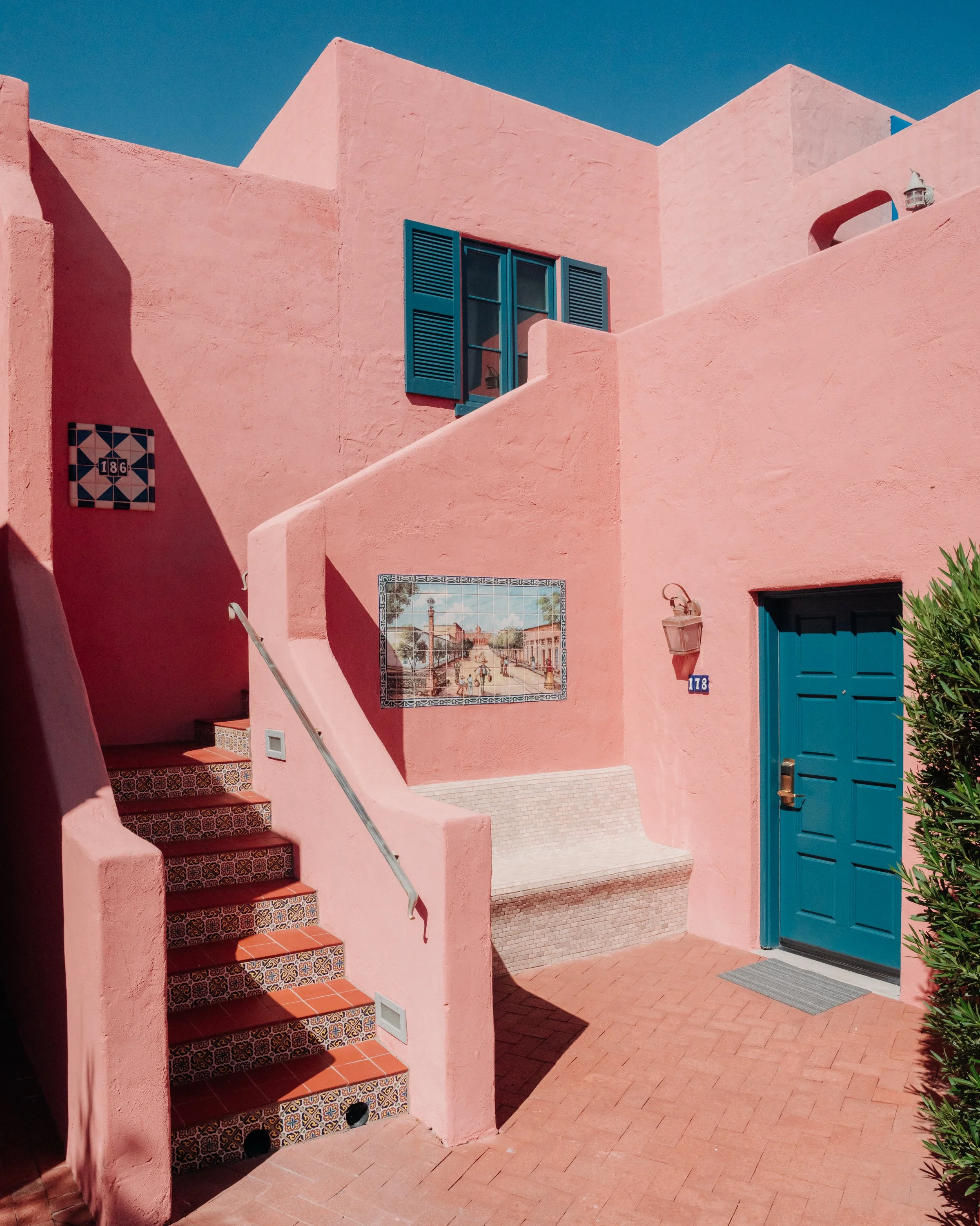 Pink building with blue window shutters, tile stairs, and a colorful mural, under a clear blue sky.