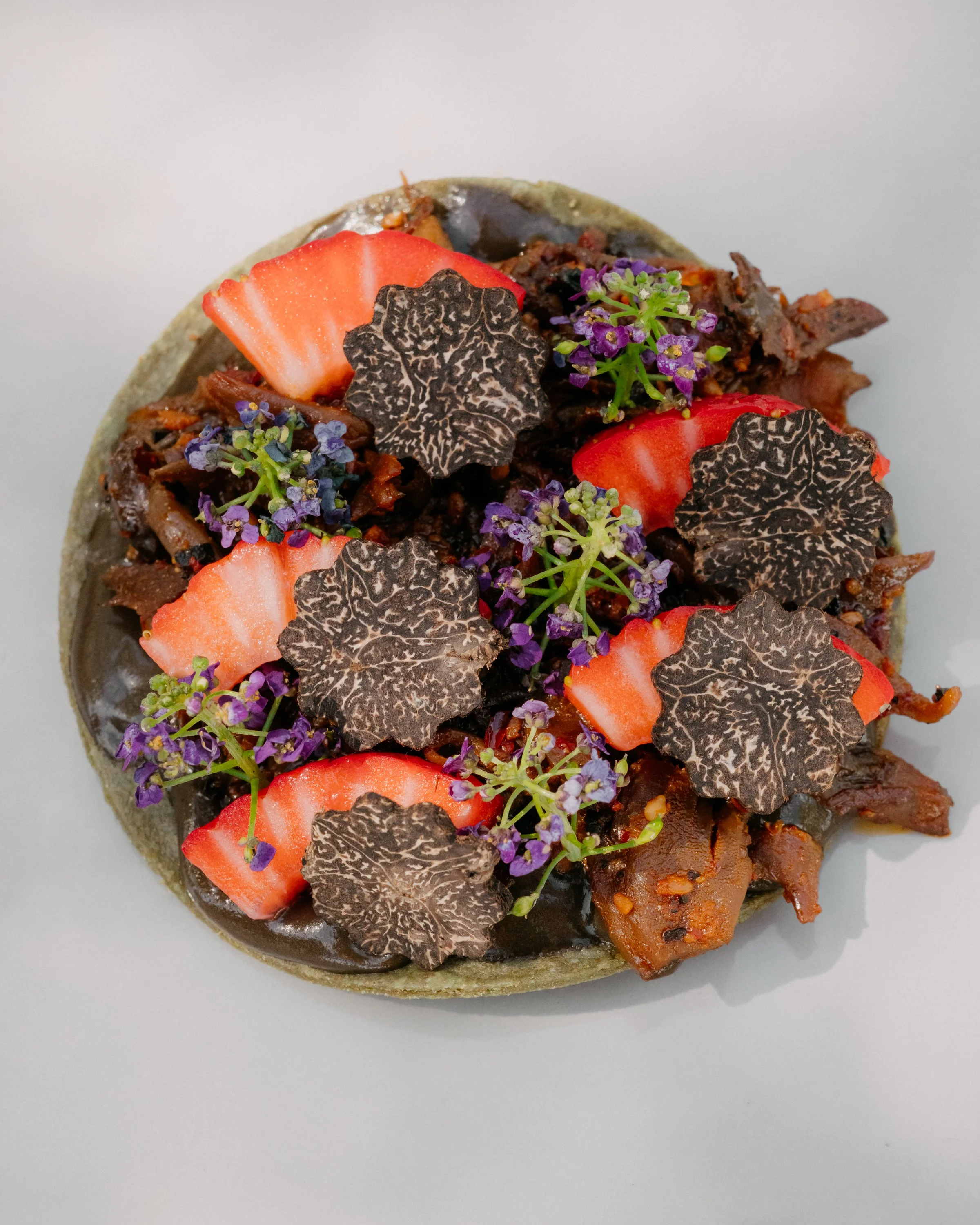 Close-up view of a tostada topped with sliced strawberries, purple edible flowers, dark decorative leaves, shredded meat, and colorful vegetables, on a white background.