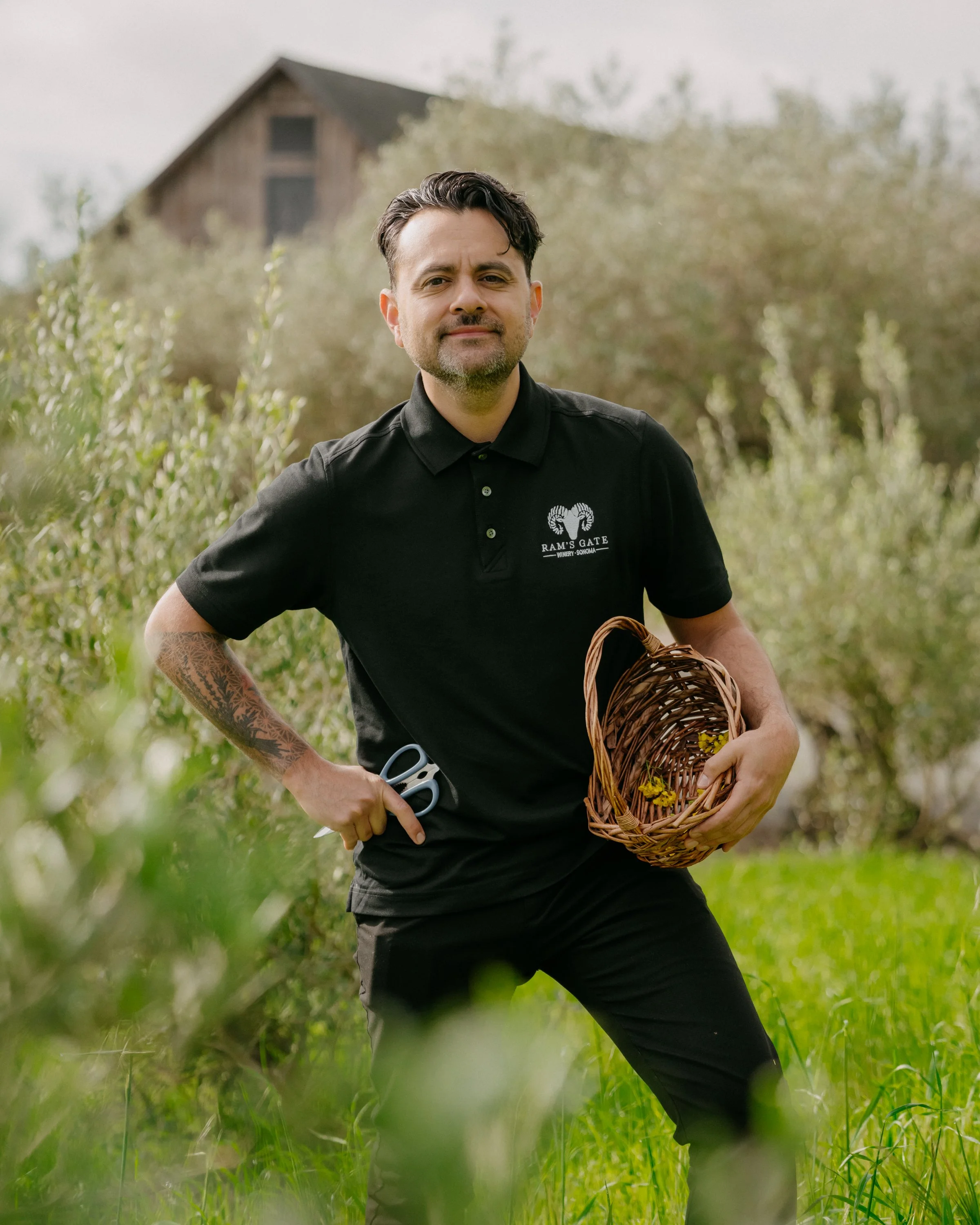 A man with dark hair, beard, and tattoos on his left arm is standing outdoors in a grassy field with trees and a barn in the background. He is wearing a black polo shirt with a ram's head logo and text. He is holding a basket with yellow flowers and 