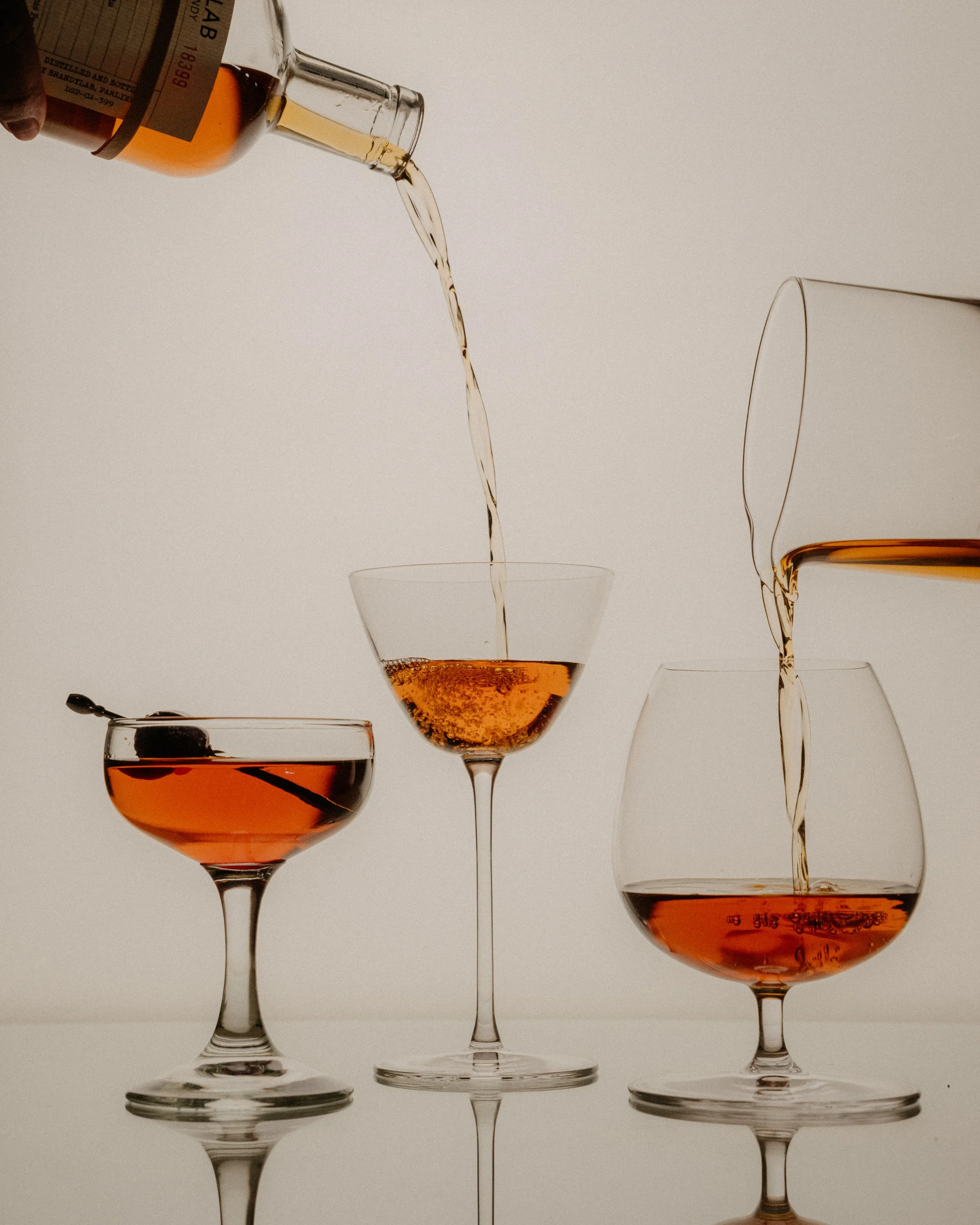 A hand pouring whiskey from a bottle into a wine glass, with other glasses on a white background.