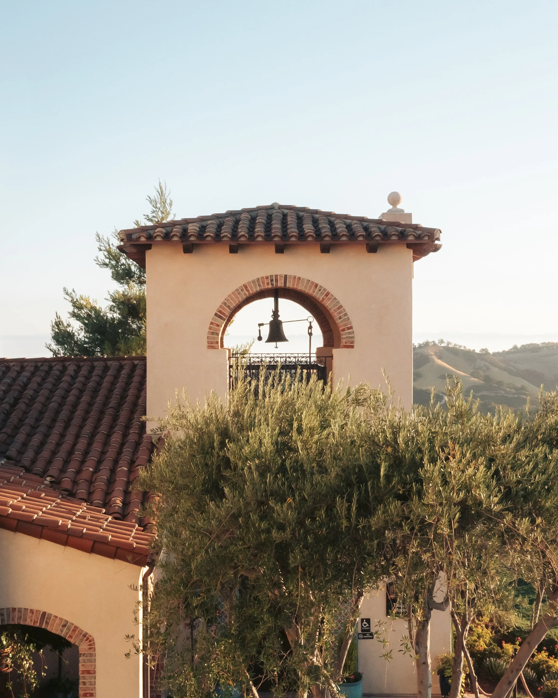 A bell tower with a bell inside, topped with a clay tile roof, and surrounded by trees and a building with arched windows and a sign indicating wheelchair accessibility.