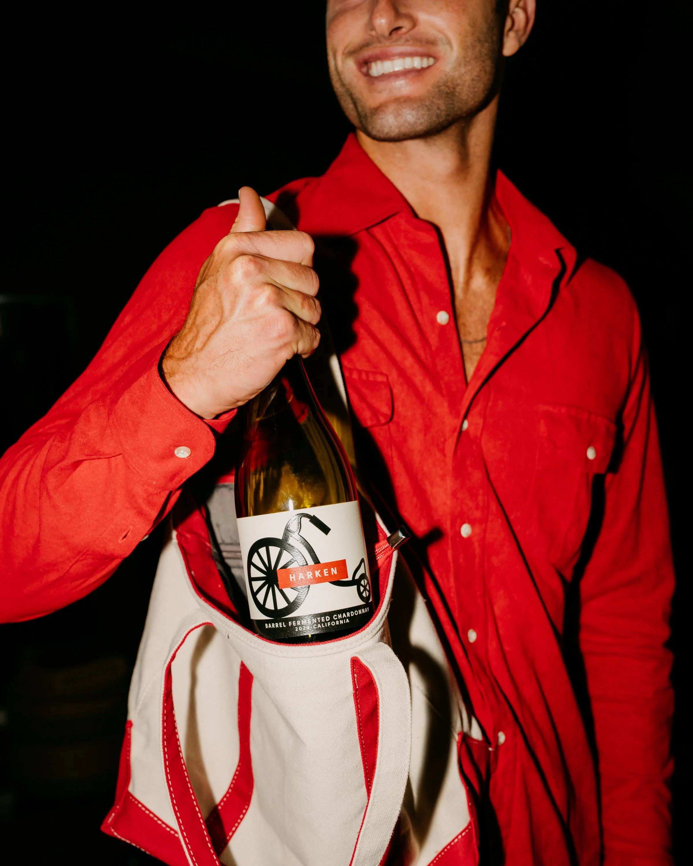 A person in a red jacket holding a bottle of wine with a label featuring a black wheel and the word 'Harken'. Only the lower part of their face is visible, with a partial smile showing white teeth, against a dark background.
