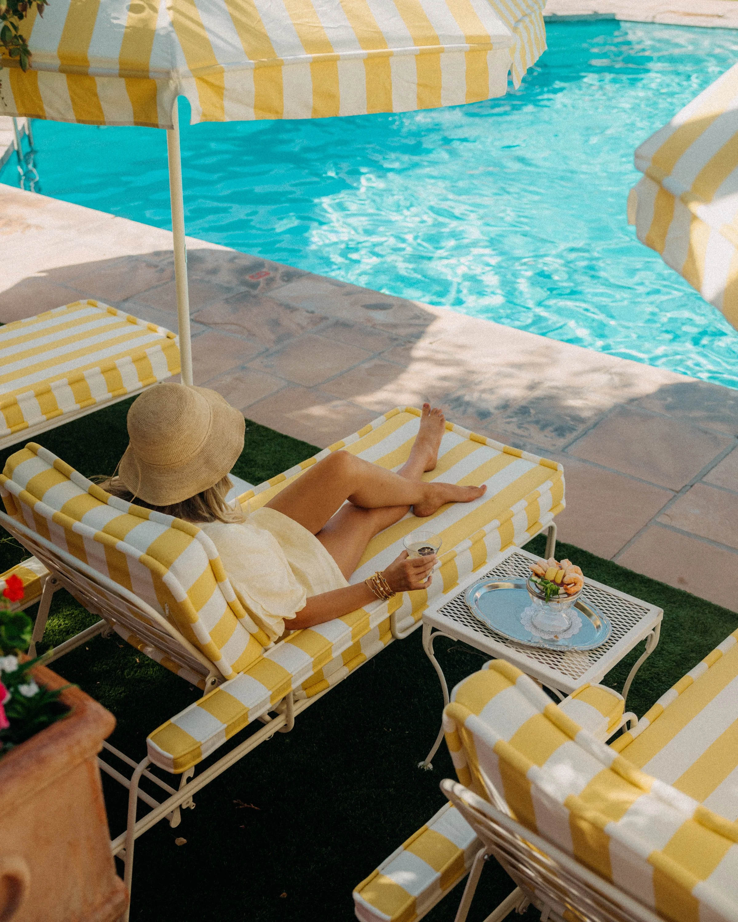 A person relaxing by a swimming pool on a yellow and white striped lounge chair, holding a wine glass, with a tray of snacks nearby, shaded by yellow and white striped umbrellas.