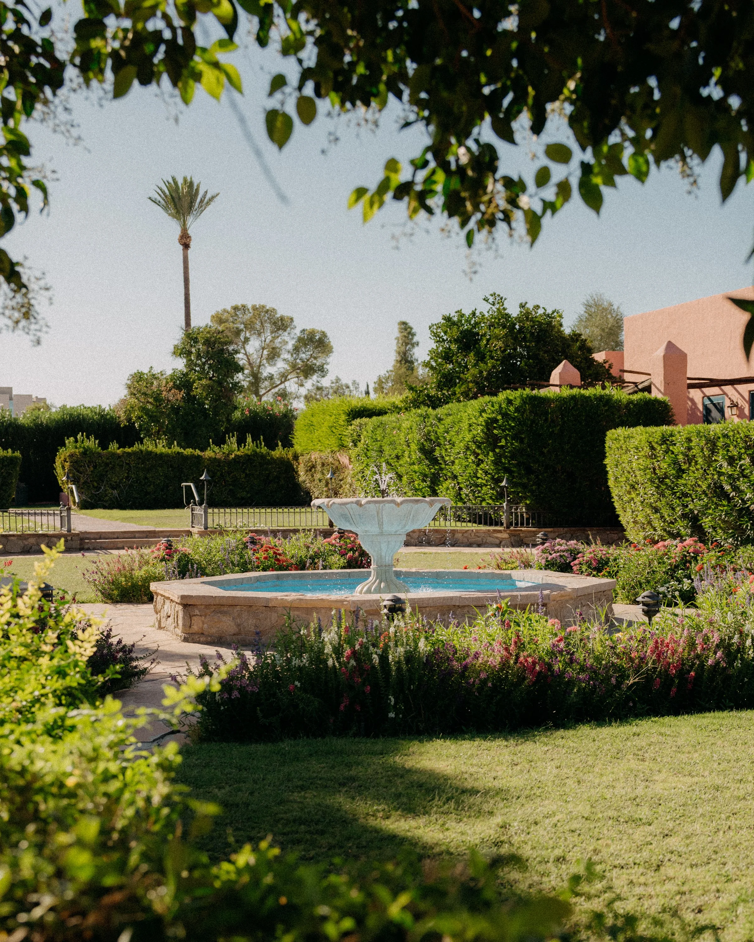 A well-maintained garden featuring a white fountain, lush green bushes, colorful flowers, and a pink building in the background, under a clear sky.
