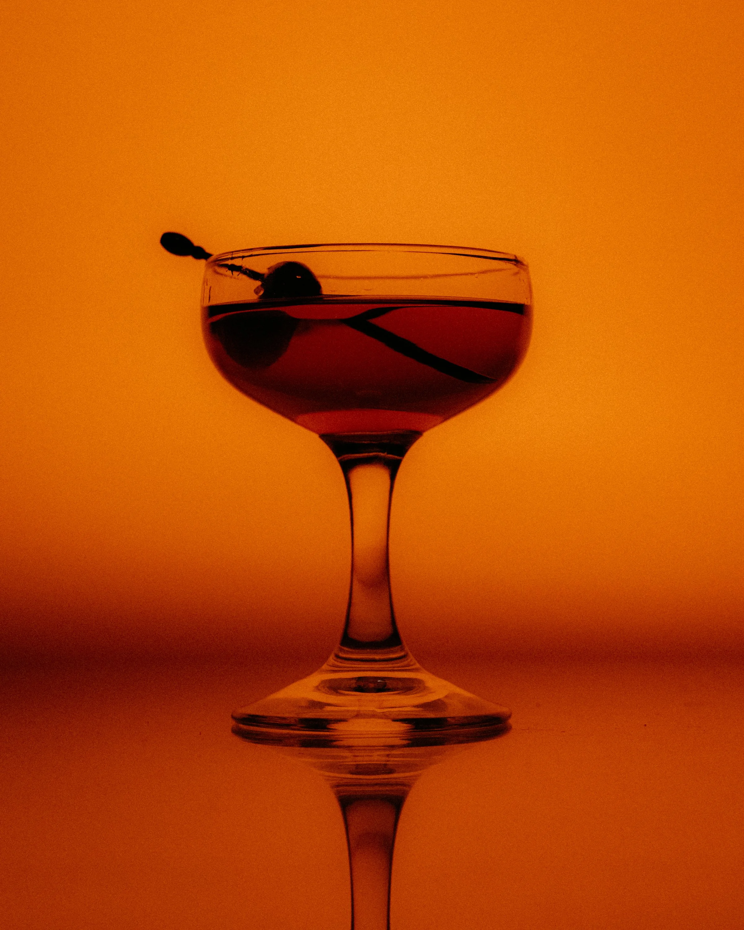 Silhouette of a cocktail glass filled with a dark red beverage, garnished with a cherry and a maraschino cherry, against an orange gradient background.
